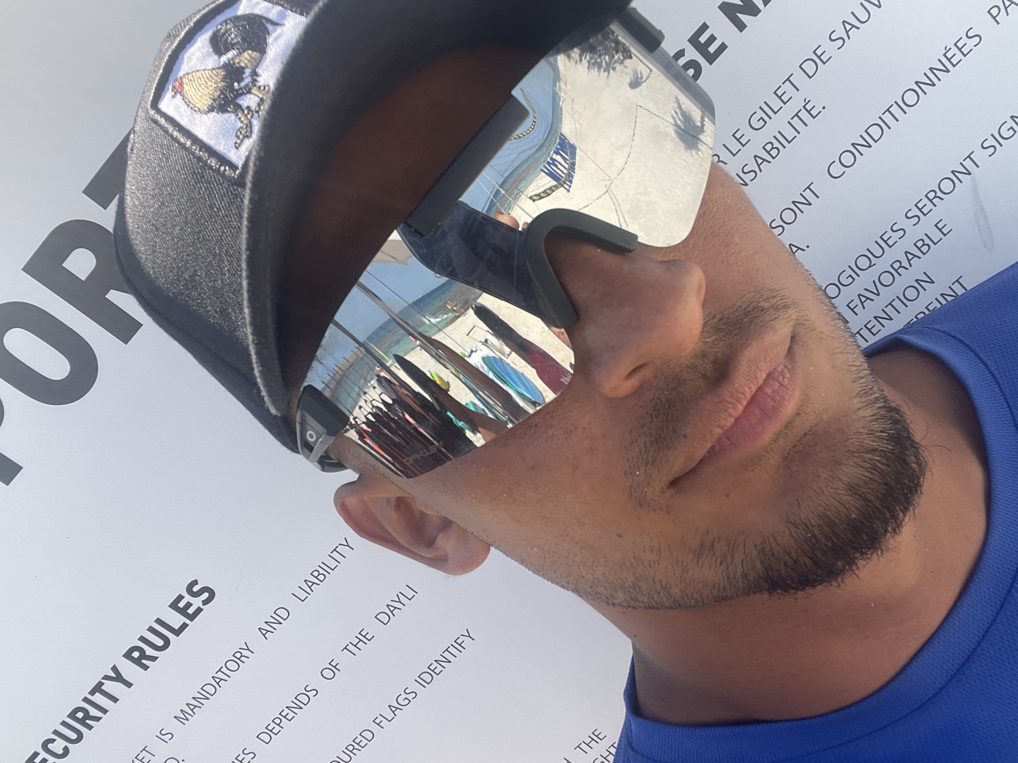 A close-up shot shows a person wearing mirrored sunglasses and a black baseball cap with an emblem. The sunglasses reflect a beach scene with surfboards and people. The person is wearing a blue shirt. The background features a white sign with the text "SECURITY RULES" and additional text in both English and French, indicating safety guidelines. The sign reads, in part, "LE GILET DE SAUV" and "SONT CONDITIONNÉES PA," suggesting the location is a public beach with lifeguards. The presence of both English and French text suggests a tourist area. Based on the reflection in the sunglasses, the surfboards, and the signage, the location is likely a beach in Playa del Carmen, Mexico.