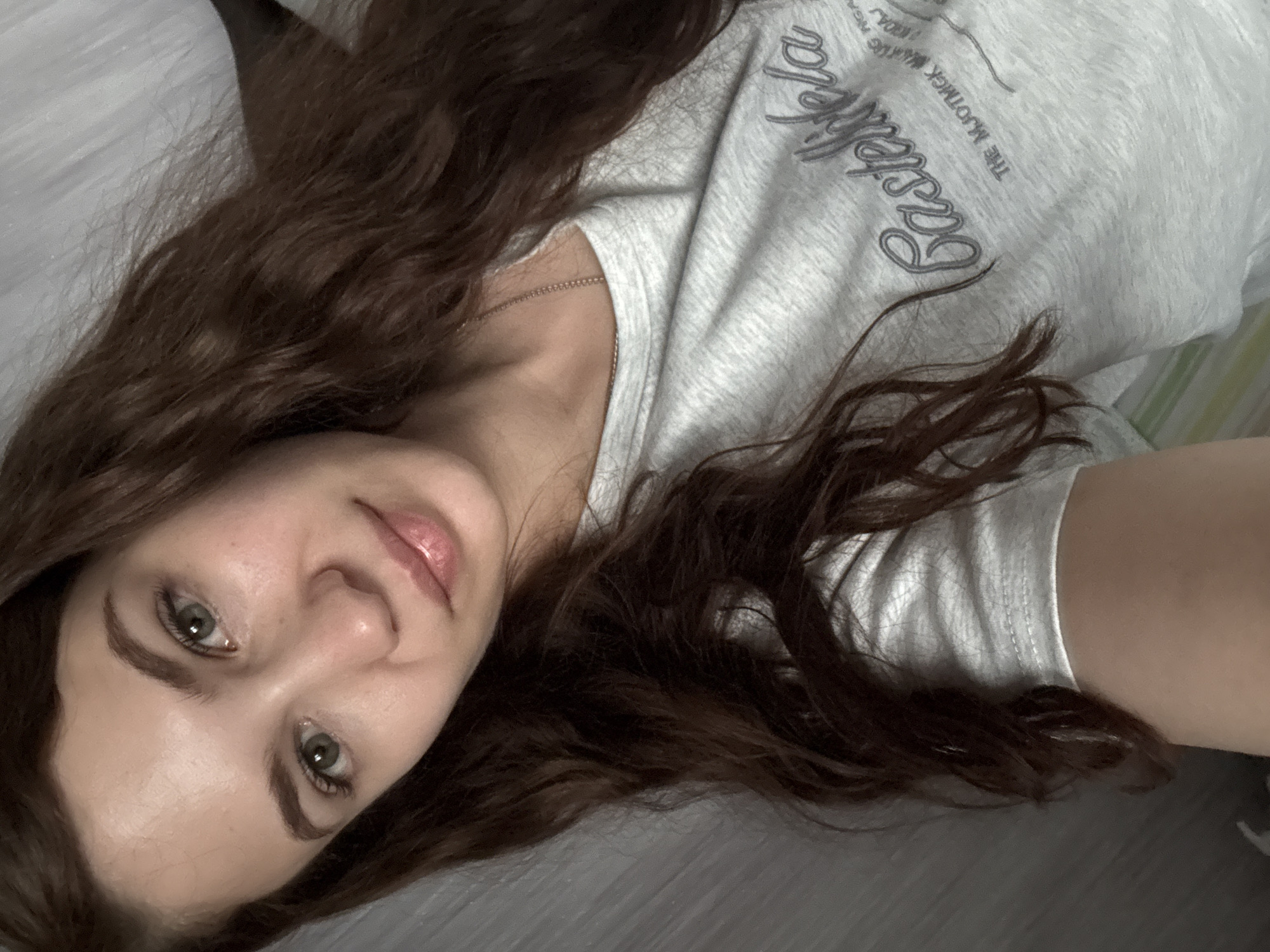 A young woman lies on what appears to be a gray surface, possibly a bed or floor. She has long, wavy brown hair that is spread around her head. She is wearing a light gray t-shirt with a logo on the upper right chest that reads, "East Life." She is looking directly into the camera. A thin silver necklace is visible around her neck. The environment gives no clear indication of being in Nizhnyaya Gar', Russia.