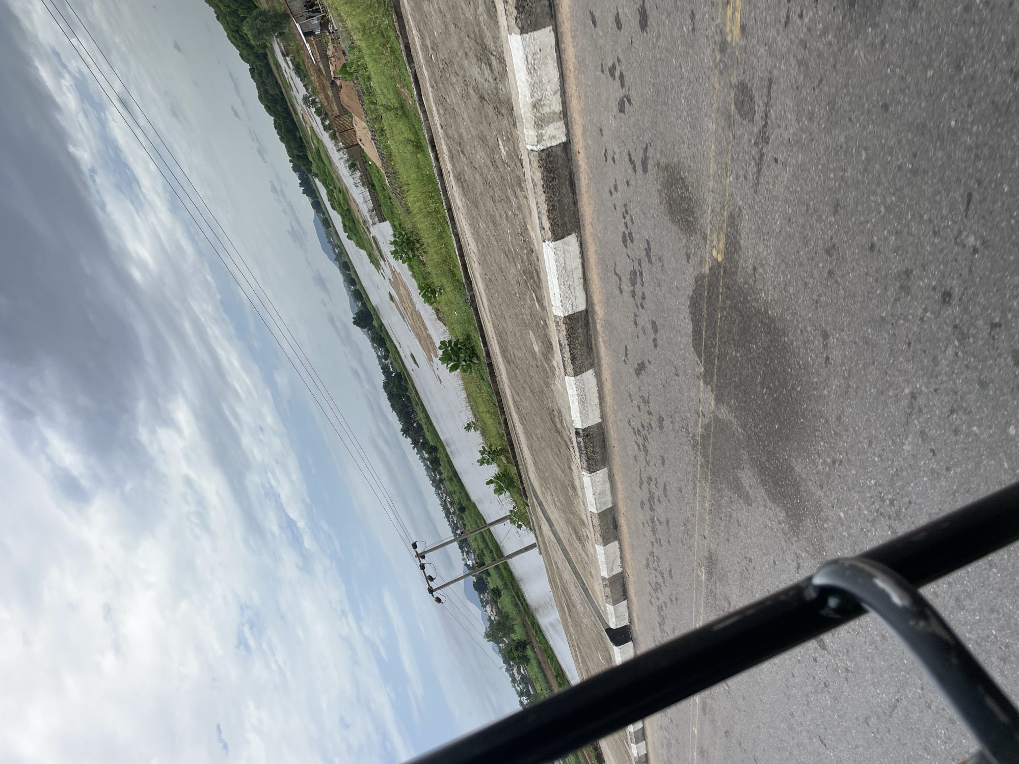 The image depicts a roadside scene, likely in Jalingo, Nigeria. The perspective is from a vehicle, with a black metal railing in the foreground. The road itself is asphalt, with visible yellow lane markings and some dark splatters, possibly oil or water stains. Adjacent to the road is a raised concrete curb with alternating black and white markings.
To the left of the road, a body of water stretches into the distance, reflecting the overcast sky. The water appears to be shallow in some areas, with patches of exposed earth and vegetation along its banks. Green grass grows alongside the water and the road, with a few scattered trees and some small bushes. In the background, there are hints of buildings and structures, suggesting a semi-urban or developing area. Power lines supported by utility poles are visible crossing the scene.
The sky is filled with a thick layer of clouds, indicating an overcast or possibly rainy day. The light is diffused, and there are no strong shadows, suggesting it is likely daytime, perhaps late morning or early afternoon. The overall mood is somewhat subdued due to the cloudy weather. No people are visible in the frame. The image conveys a sense of everyday life in a location where water and infrastructure coexist. There is no visible text in the image.