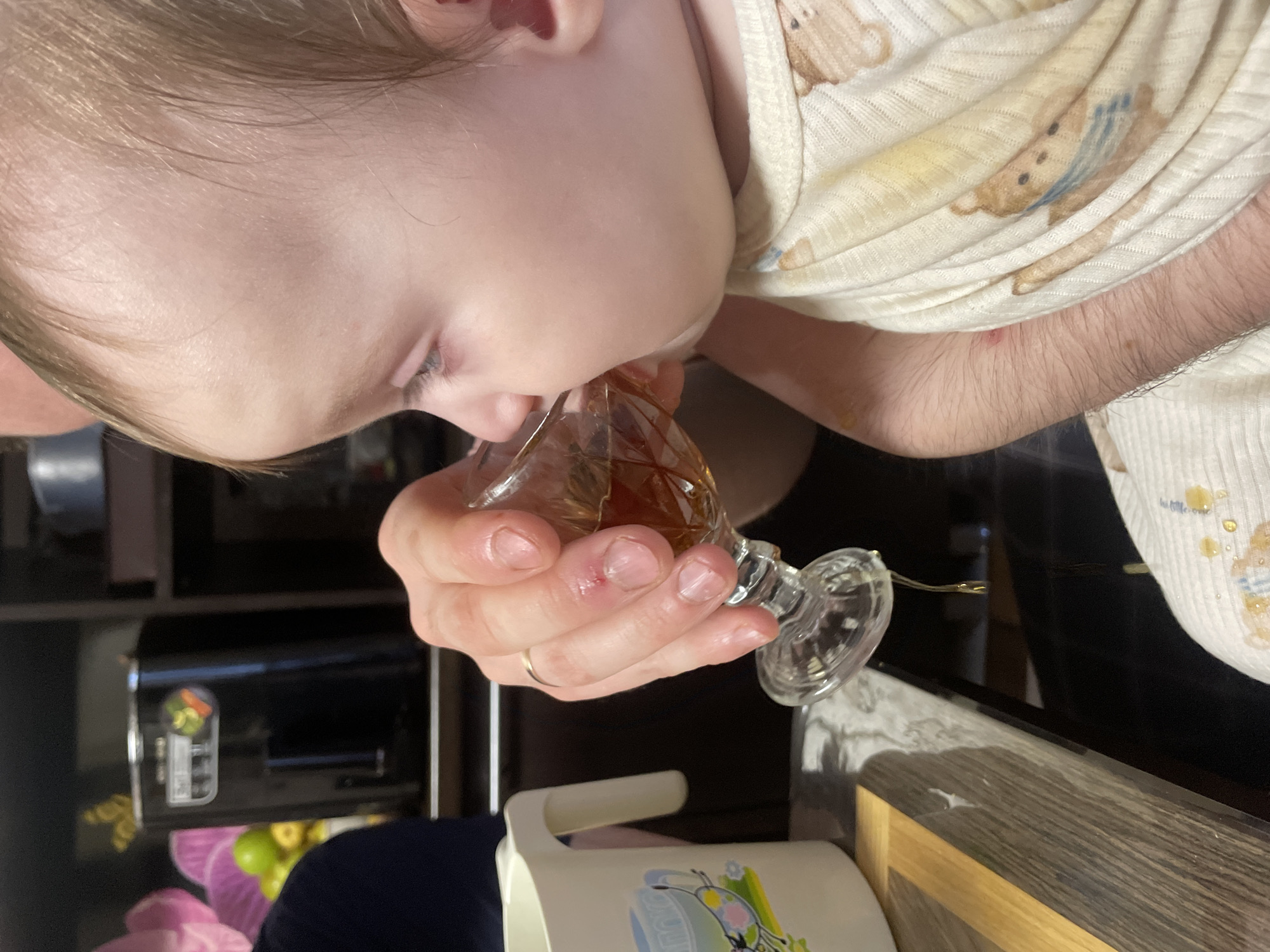 The image displays a close-up, rotated view of a baby or young child drinking from a small, faceted glass held by an adult's hand. The child, with light hair and fair skin, is wearing a light-colored top with teddy bear patterns. Their mouth is on the rim of the glass, consuming an amber-colored liquid, likely a non-alcoholic beverage such as juice or tea.
The adult's hand, adorned with a simple gold band on the ring finger, firmly grips the stemmed glass. A small red mark is visible on one of the adult's knuckles. Part of the adult's arm, covered in hair, extends from beneath a dark sleeve.
The setting is indoors, likely a home kitchen or dining area. In the background, a black electronic appliance, possibly a coffee maker, rests on a dark shelf, featuring a colorful label with indistinct text and icons. Below this, a white pitcher with a handle and a colorful butterfly design is partially visible. Further back, some green and yellow round objects, possibly fruits, can be seen in a container. A wooden surface or tabletop occupies the right side of the frame. The lighting suggests an indoor environment. No explicit location cues specific to Kuyeda, Russia, are discernible within the image.