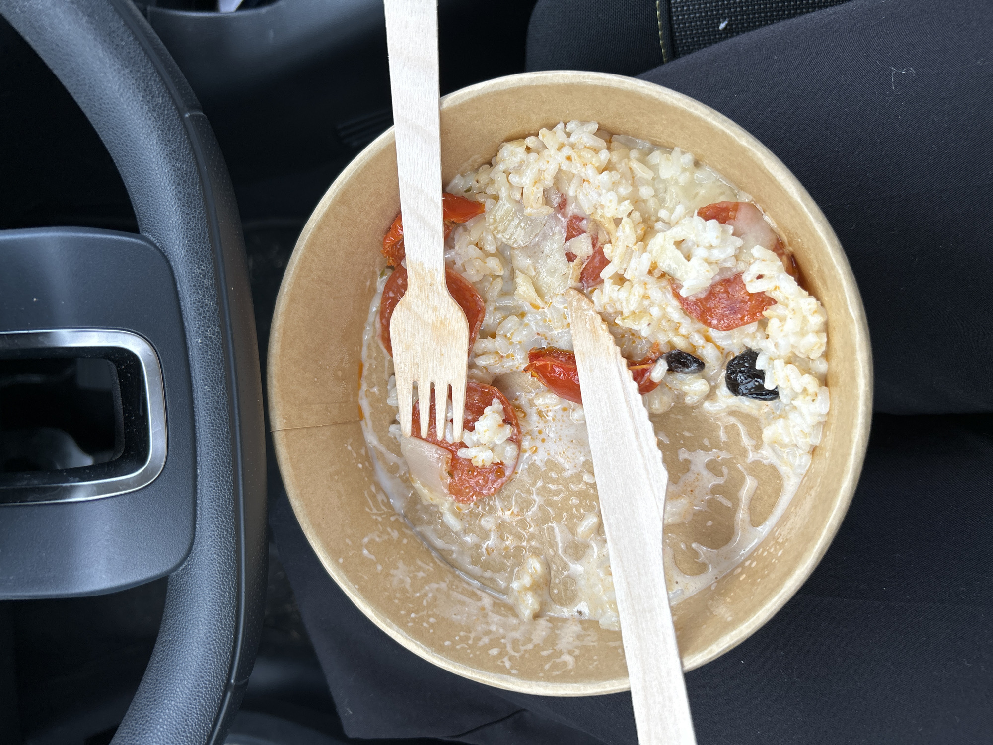 The image shows a partially eaten bowl of risotto in a brown cardboard container. There are wooden cutlery items, a fork and a knife, stuck in the food. The risotto appears to contain rice, tomatoes, olives, and cheese. The bowl is resting on black fabric, possibly clothing, inside of a car, with the steering wheel visible to the left. The location is in a car somewhere in Valence, France.