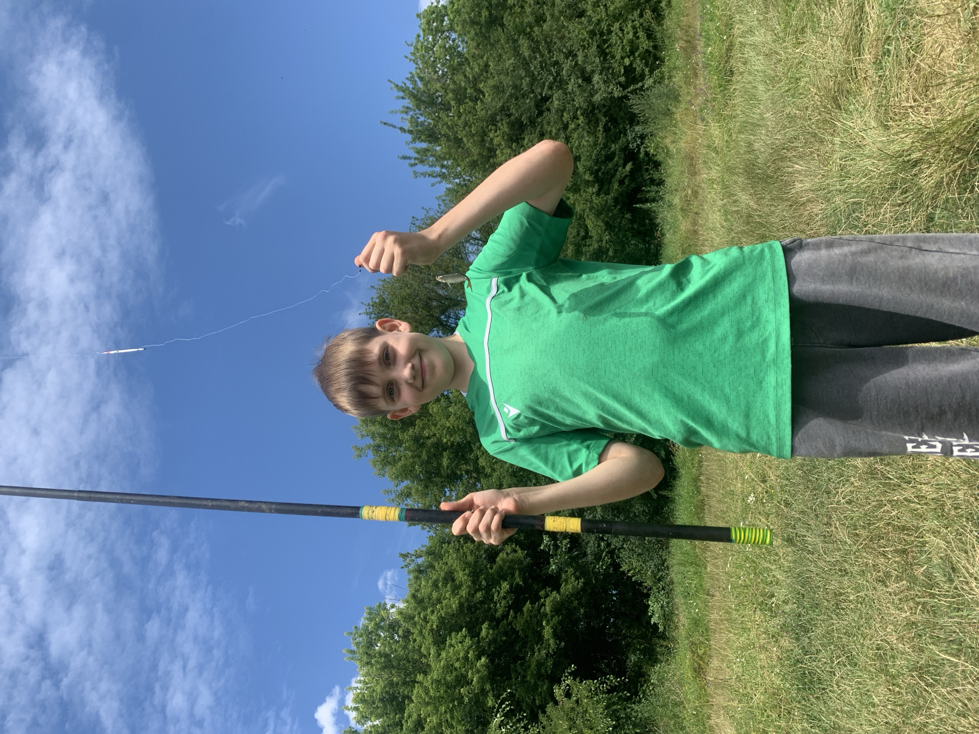 A young boy, approximately 10-12 years old, stands in a grassy field under a bright blue sky with scattered white clouds. He is holding a fishing rod with his left hand, and in his right hand, he proudly displays a small fish caught on the line. The boy is wearing a green t-shirt with white stripes on the sleeves and dark gray sweatpants. The fishing rod is tall and black with yellow and green accents near the bottom. Lush green trees form a backdrop behind him, suggesting a rural or park-like setting. The overall impression is one of a successful fishing excursion on a sunny day.