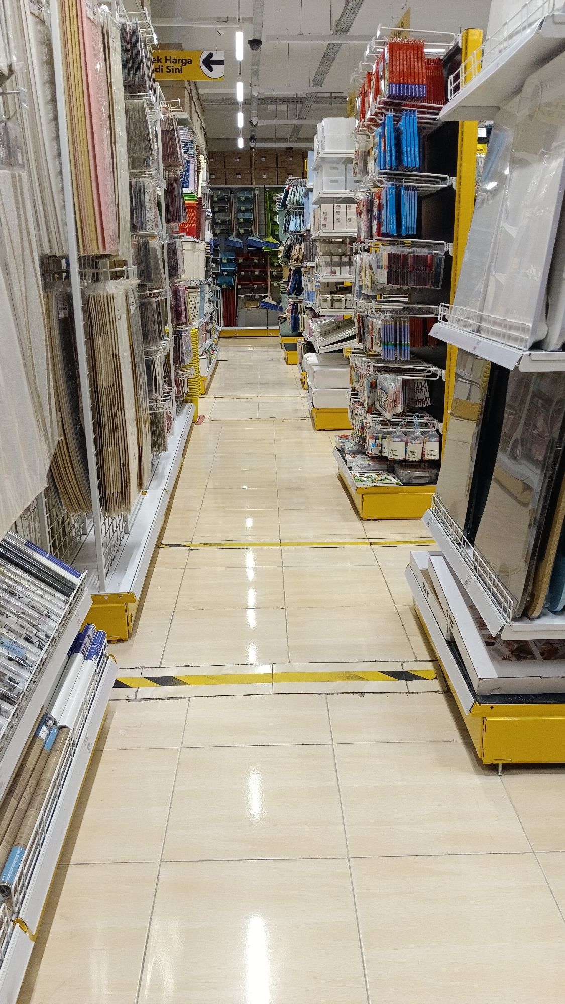 The image depicts an aisle in a retail store, likely a department store or home goods store, with shelves of merchandise lining both sides. The aisle is well-lit with overhead fluorescent lighting. The floor is tiled in a light beige color, and there are yellow and black caution stripes on the floor indicating a walkway or queue line.
On the left side of the aisle, shelves display various items, including what appear to be fabric rolls or hangings, possibly wallpaper or decorative materials, and packaged goods. On the right side, shelves hold an assortment of items, including stacks of white storage boxes, smaller packaged goods, and what look like framed items or large flat objects in clear packaging. Some of the merchandise is in plastic wrapping.
Further down the aisle, more shelves are visible, stocked with various products. In the distance, there are more shelves and boxed inventory. A sign with Indonesian text and an arrow pointing to the left is visible in the upper left portion of the image.
The environment appears clean and organized, typical of a retail space. There are no people visible in the image. The overall impression is a commercial setting designed for customer browsing and purchasing. The specific location of Gunting Saga, Indonesia, is not directly inferable from the visual content of the image itself, but the presence of Indonesian text on the sign suggests it is located in Indonesia.