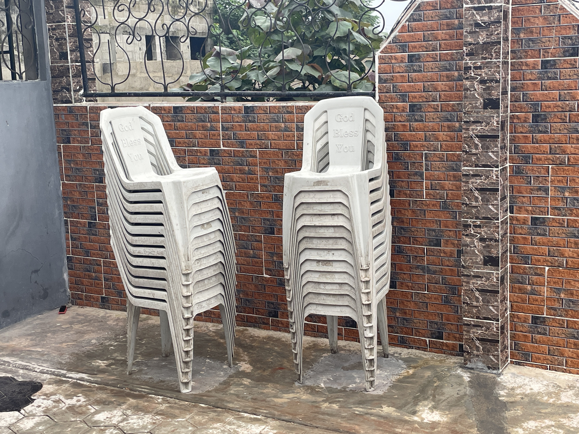 Two stacks of white plastic chairs are positioned on a wet, concrete patio in Iyafin, Nigeria. The chairs are stacked high, with the top chair in each stack bearing the embossed inscription "God Bless You." Behind the chairs, a brick wall features a patterned tile accent running vertically. The wall is partially obscured by a dark green, leafy plant that grows behind a black metal railing. To the left of the chairs, a gray, concrete wall and a portion of a building with arched windows are visible. The ground is wet and reflects the surrounding structures. A small, red object, possibly a cellphone, is situated on the ground near the left wall.