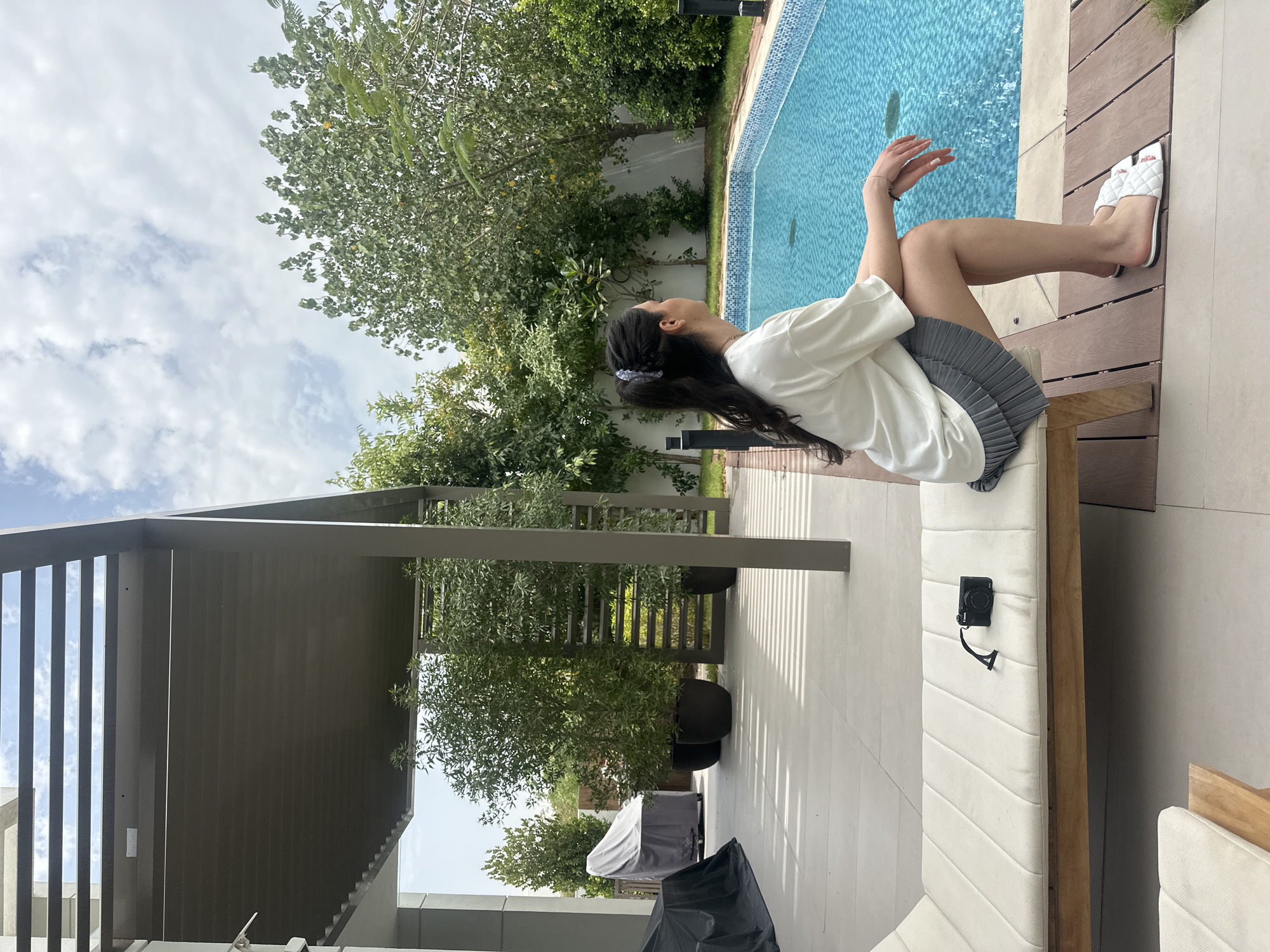 This image depicts an outdoor leisure scene at a residence or resort in Dubai, United Arab Emirates, on a partly cloudy day. A female individual, seen from a side-back angle, is the central subject. She has long, dark hair secured with a light-colored clip and is dressed in a loose white t-shirt and a grey pleated skirt. She wears white quilted slides on her feet. The woman is seated on a white cushioned outdoor bench, her gaze directed towards a large, rectangular swimming pool with bright blue tiling. Her right hand is gently raised, suggesting a moment of relaxation or contemplation.
The setting is a well-appointed patio area featuring a swimming pool, surrounded by modern light-colored paving and wooden decking. Lush green trees and dense foliage form a natural backdrop around the property, contributing to a sense of privacy. To the left, a contemporary outdoor structure, likely a pergola or patio roof, with dark grey slatted elements, provides shade over part of the outdoor living space. Beneath this structure, several trees are planted in large black pots, and two covered outdoor items, possibly grills or furniture, are visible. The overall atmosphere is serene and suggestive of a comfortable outdoor living space. The weather appears pleasant with visible white clouds against a bright sky. There is a black compact camera with a strap resting on the bench next to the person. No discernable text is visible in the image.