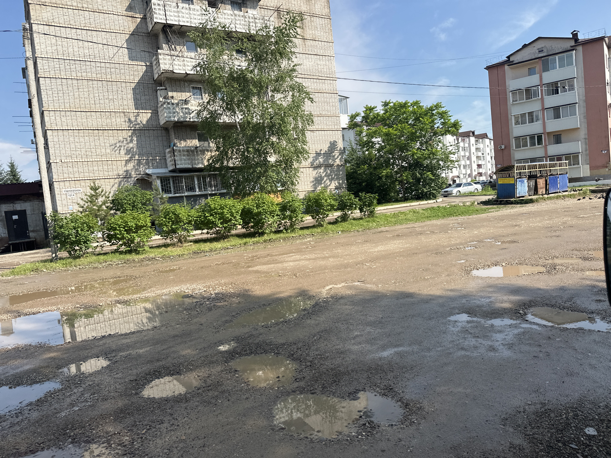 This media file depicts an outdoor scene in Birobidzhan, Russia, likely during the daytime under a bright, partly cloudy sky. The primary focus is a weathered, unpaved road in the foreground, heavily damaged with numerous potholes filled with muddy water, reflecting the surrounding environment. To the left, a tall, multi-story apartment building constructed with light-colored brick dominates the frame. It features multiple balconies with visible railings and a somewhat worn appearance. Adjacent to the building is a row of neatly trimmed green bushes.
Behind the apartment building and bushes, several trees are visible, including a prominent birch tree with a slender trunk and dense green foliage. Further in the background, another modern apartment building with a pink and white facade stands. Near this building, a cluster of blue and brown trash bins are situated, suggesting a residential area. A silver car is parked on the road near the trash bins. The overall setting appears to be a typical urban or suburban residential area in Russia, characterized by aging infrastructure and greenery. There is no discernible activity or event taking place, and the scene conveys a sense of ordinary daily life. No specific emotions are evoked, other than perhaps a mild impression of neglect due to the road's condition. The visible text is limited to a sign on the brick apartment building, which reads "ул. Школьная, 121-6".