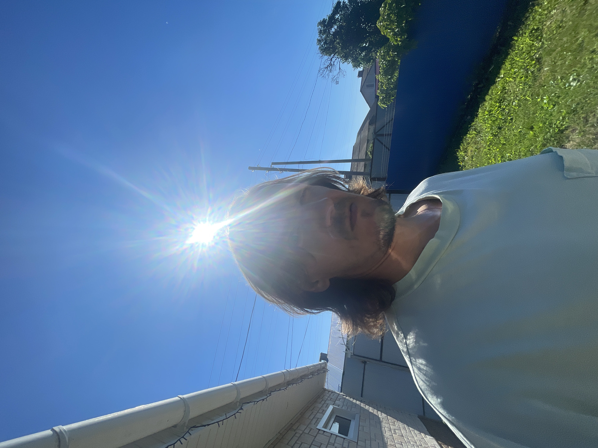 A person is standing outdoors in front of a building on a sunny day. The person is wearing a light blue t-shirt and has medium-length brown hair. The sun is shining brightly in the sky, causing lens flare. In the background, there is a blue fence, some trees, and houses. The area appears to be a residential neighborhood.