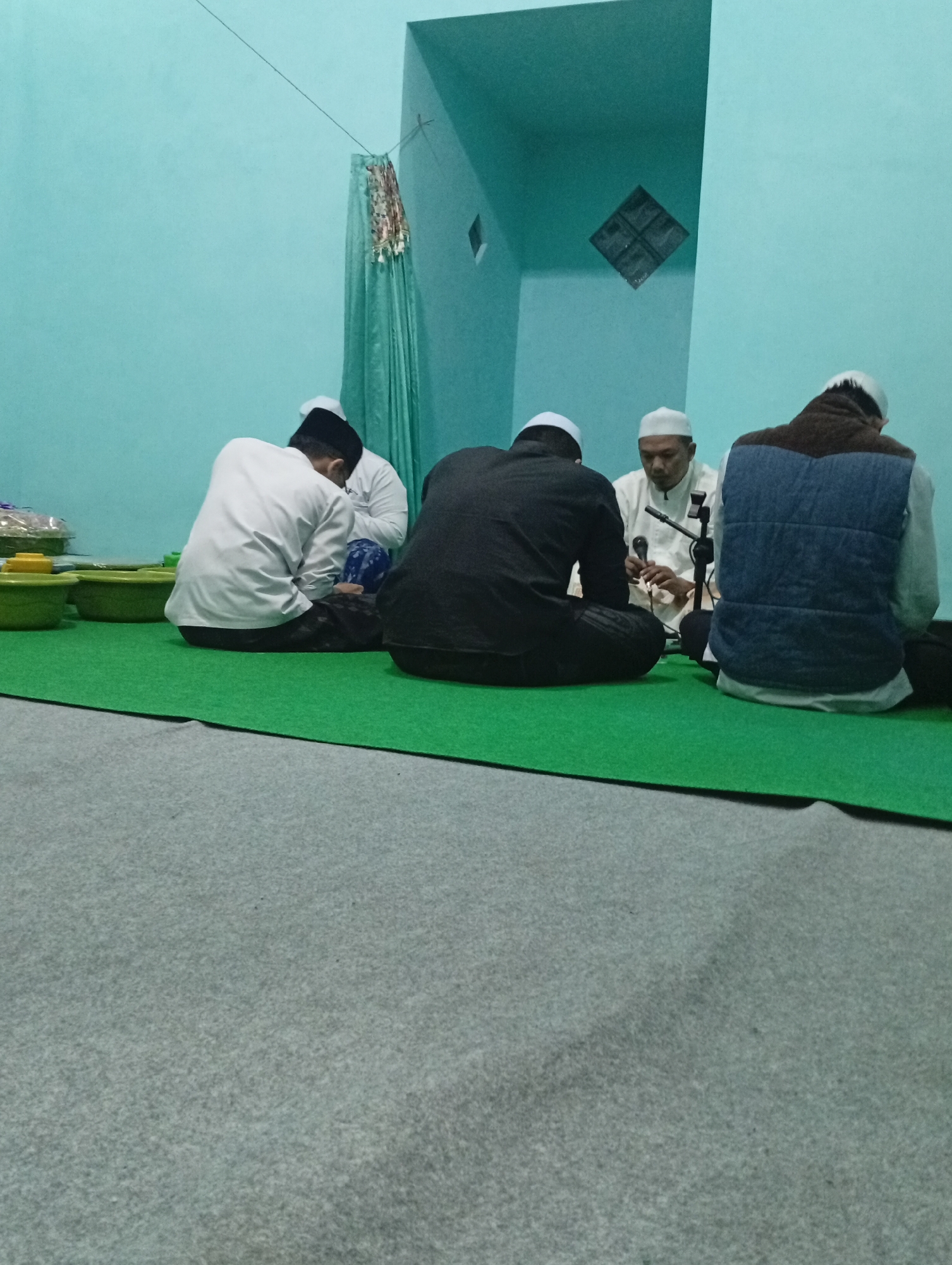 In Jatisari, Indonesia, five individuals are seated on a green carpeted floor within a room with light blue walls. One individual, facing away from the camera, wears a white long-sleeved shirt and a black skullcap, seated with legs crossed. To their left, another individual, also facing away, wears a black long-sleeved garment and a white skullcap. A third individual, in the center and facing the camera, wears a white long-sleeved shirt and a white skullcap, holding a microphone attached to a stand. Another person, on the right, is seen from the back wearing a blue puffer vest over a brown garment and a white skullcap. The background features a green curtain hanging to the left and a decorative element on the ceiling. Several green containers are placed on the floor to the left.