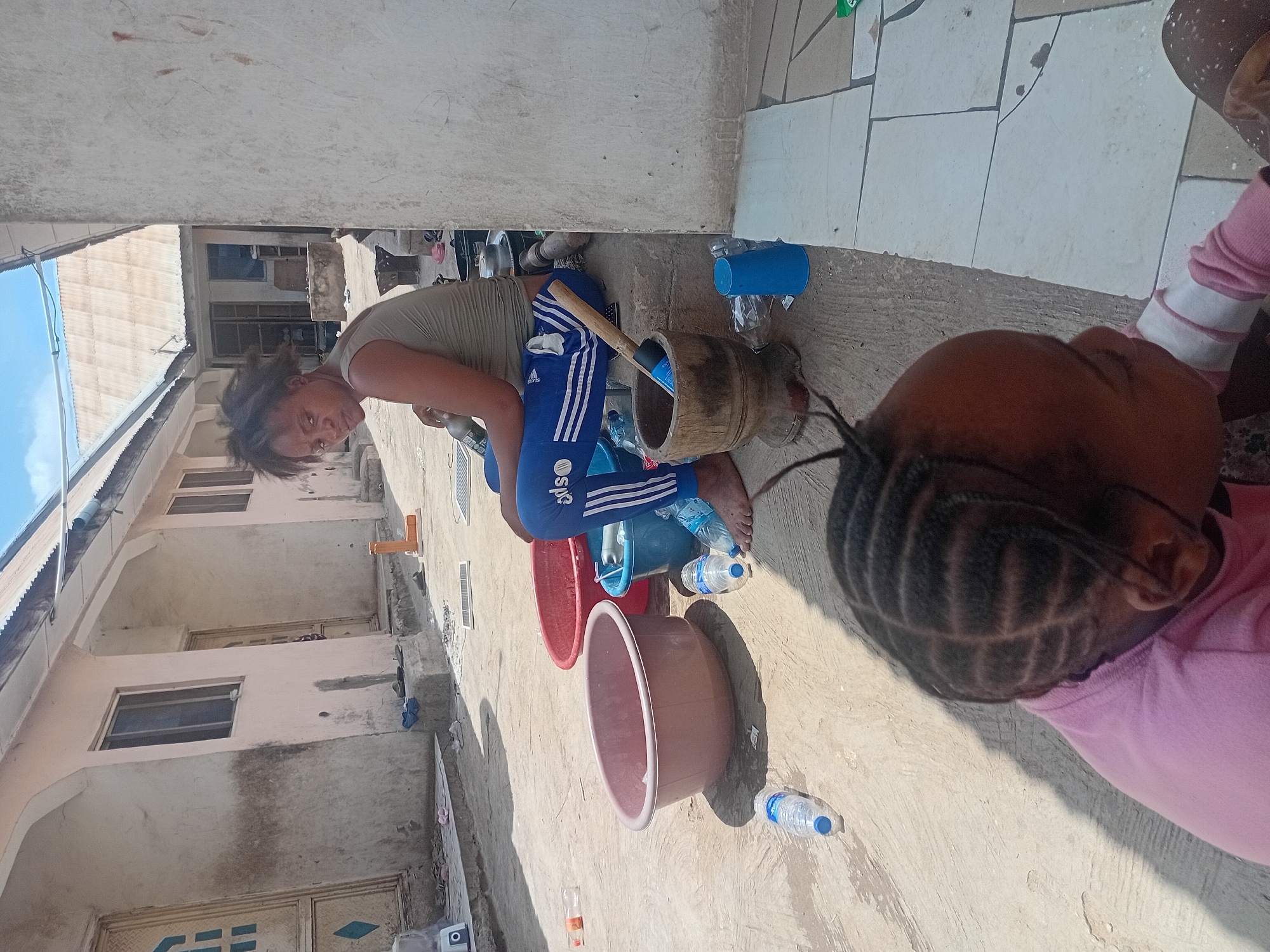 The image captures an outdoor scene, likely a courtyard or a paved area outside a residential building, under bright daylight in Lokoja, Nigeria.
In the center, a woman is squatting or kneeling on a concrete ground. She wears a greenish-brown short-sleeved top and blue Adidas track pants with white stripes, featuring the "Adidas" logo and "SOS" text on the leg. Her dark hair is somewhat disheveled, and she looks directly at the camera with a neutral expression. Next to her is a wooden mortar with a wooden pestle inside, suggesting food preparation or processing. Several plastic containers are scattered around her, including red, brown, and blue basins or buckets, some containing water or other liquids. Multiple clear plastic water bottles, some bearing the "NESTLE Pure Life" label, are also on the ground. A blue plastic cup is visible nearby.
In the immediate foreground, partially obscuring the lower right, is the back of a child's head and shoulder. The child is wearing a pink top, and her dark hair is styled in neat cornrows. She is looking away from the camera.
The background shows parts of a multi-story building with a light-colored facade, visible windows, and metal grates. Corrugated metal roofing is visible further back under a clear blue sky, indicating sunny weather. The ground beneath the individuals is a mix of rough concrete and light-colored square tiles, with strong shadows suggesting the sun is high. The overall scene depicts an everyday activity in an outdoor residential setting.