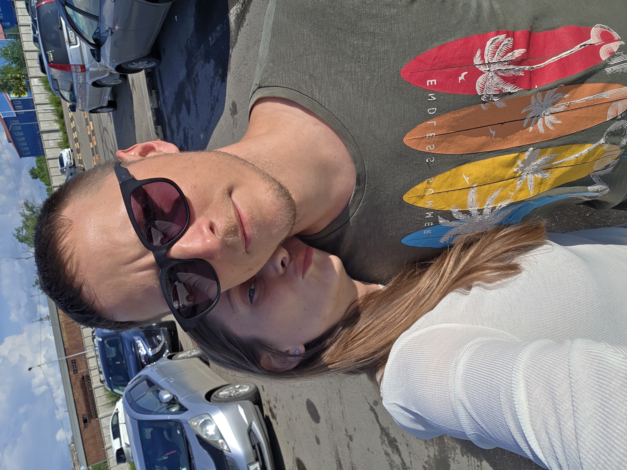 This is a candid, eye-level, selfie-style photo taken outdoors in a parking lot. A man and a woman are the primary subjects, positioned in the foreground. The man, wearing sunglasses and a dark green t-shirt with a graphic print of surfboards and palm trees, is looking towards the camera, with his left shoulder slightly turned. A woman is leaning her head on his shoulder and has her eyes closed and lips puckered, as if about to kiss him. She is wearing a white ribbed long-sleeved top. Several cars are visible in the background, including a silver hatchback, a dark SUV, and other vehicles parked in rows. The environment suggests an urban setting with paved ground and some industrial-looking buildings in the distance under a partly cloudy sky. The location is identified as a parking lot in Birobidzhan, Russia, based on context.