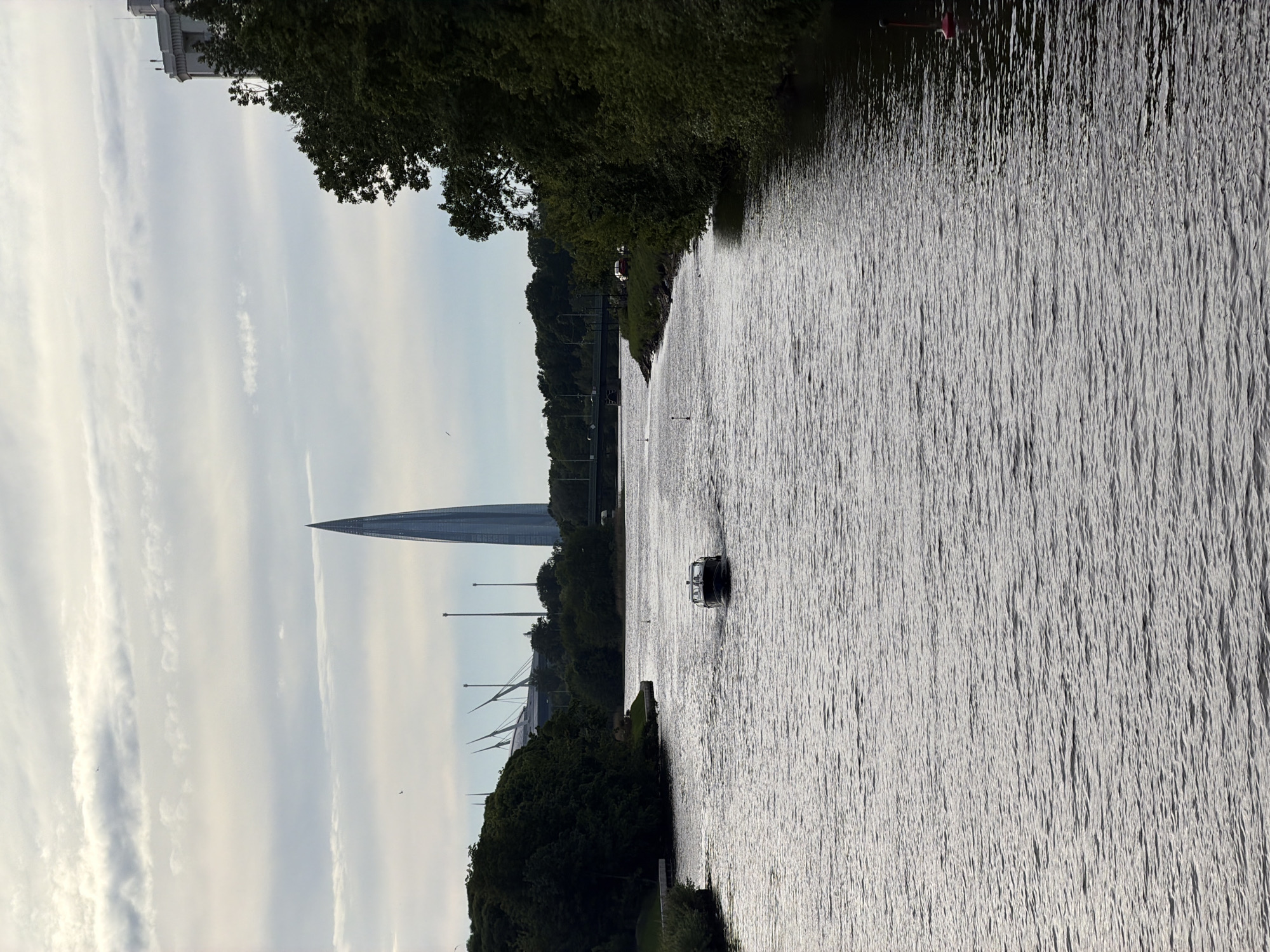 The image captures a scenic view of a wide river or canal in Saint Petersburg, Russia, bordered by lush green banks, under a bright sky. The perspective appears to be from a bridge or an elevated viewpoint.
On the left side of the foreground, dense green trees line the bank. The river itself dominates the lower and central parts of the frame, displaying numerous ripples that catch and reflect the sunlight, creating a shimmering effect across its surface. A dark-colored motorboat is visible on the water, cruising from the right side towards the left, leaving a subtle wake behind it.
In the background, on the far bank, a distinctive and very tall modern skyscraper, identified as the Lakhta Center, rises prominently on the right, its pointed top piercing the sky. To the left of the skyscraper, other large, modern architectural structures with complex cable-stayed designs, consistent with parts of the Gazprom Arena (Krestovsky Stadium), are visible amidst the trees.
The sky in the upper portion of the image is light-colored, suggesting either a bright overcast day or late afternoon/early evening light. Several elongated, wispy white clouds stretch across the expanse of the sky. The overall lighting indicates a calm, clear day. No people are explicitly visible, and there is no discernible text within the image. The scene evokes a tranquil atmosphere, blending modern urban landmarks with natural waterways and greenery.