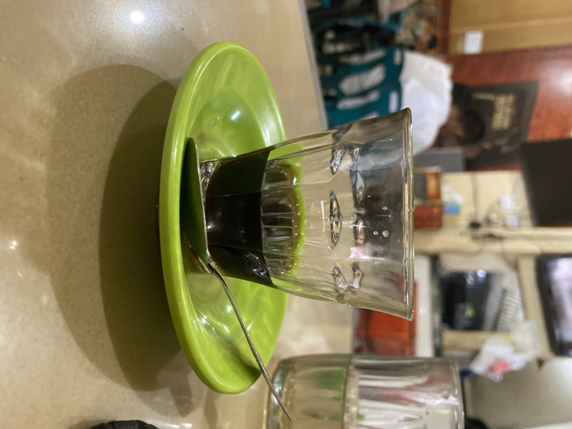 A close-up shot captures a small glass of dark liquid, likely coffee, resting on a bright green saucer. A silver spoon lies on the saucer, partially submerged in the liquid. The glass itself is clear and textured, with a slightly ribbed design. In the background, out of focus, are hints of a bustling indoor setting, possibly a cafe or restaurant in Lhokseumawe, Indonesia, with patrons and tables visible. The overall lighting suggests an indoor environment, perhaps with ambient lighting from above. The table surface appears to be a light-colored, textured material, casting a soft shadow from the glass and saucer.