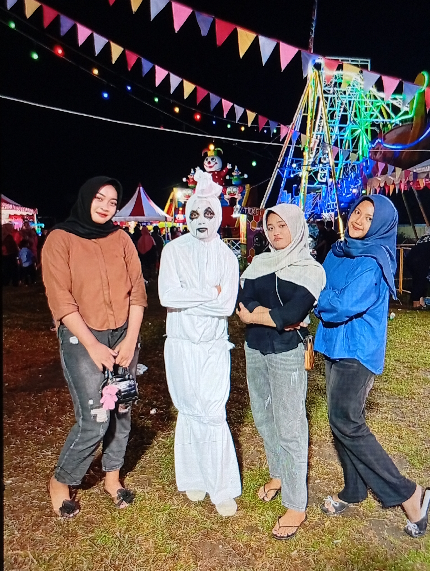 An outdoor night scene in the city of Lhokseumawe, Indonesia, captures four individuals posing for a photograph at a fair or carnival. The ground is composed of sparse, dry grass and patches of visible soil. In the background, two parallel lines of multicolored triangular bunting (red, pink, blue, yellow, white) are strung across the dark sky, accompanied by lines of small, illuminated lights in various colors. Prominently displayed on the right is a large, brightly lit fair ride with blue and green illumination. Smaller tents, one with red and white stripes, and indistinct figures of other fair attendees are visible in the mid-ground.
From left to right, the individuals are:
1. A female wearing a black hijab, a brown long-sleeved button-up shirt, and dark, ripped jeans. She holds a small black handbag with a pink teddy bear charm and wears black slip-on sandals adorned with dark floral details. She is smiling and looking directly at the camera.
2. An individual dressed in a white, full-body costume resembling a ghost or mummy, featuring a hooded headpiece and a face painted white with dark circles around the eyes and a dark mouth. Their arms are crossed over the chest, and they look forward. White footwear or foot coverings are visible.
3. A female wearing a light grey hijab, a black long-sleeved top, and light grey ripped jeans. Her arms are crossed over her chest, and she exhibits a neutral expression, looking slightly to her right. She wears black flip-flops.
4. A female wearing a blue hijab, a bright blue long-sleeved shirt, and dark trousers. Her arms are crossed over her chest, and she is smiling and looking at the camera. She wears black slip-on shoes with a white bow embellishment.