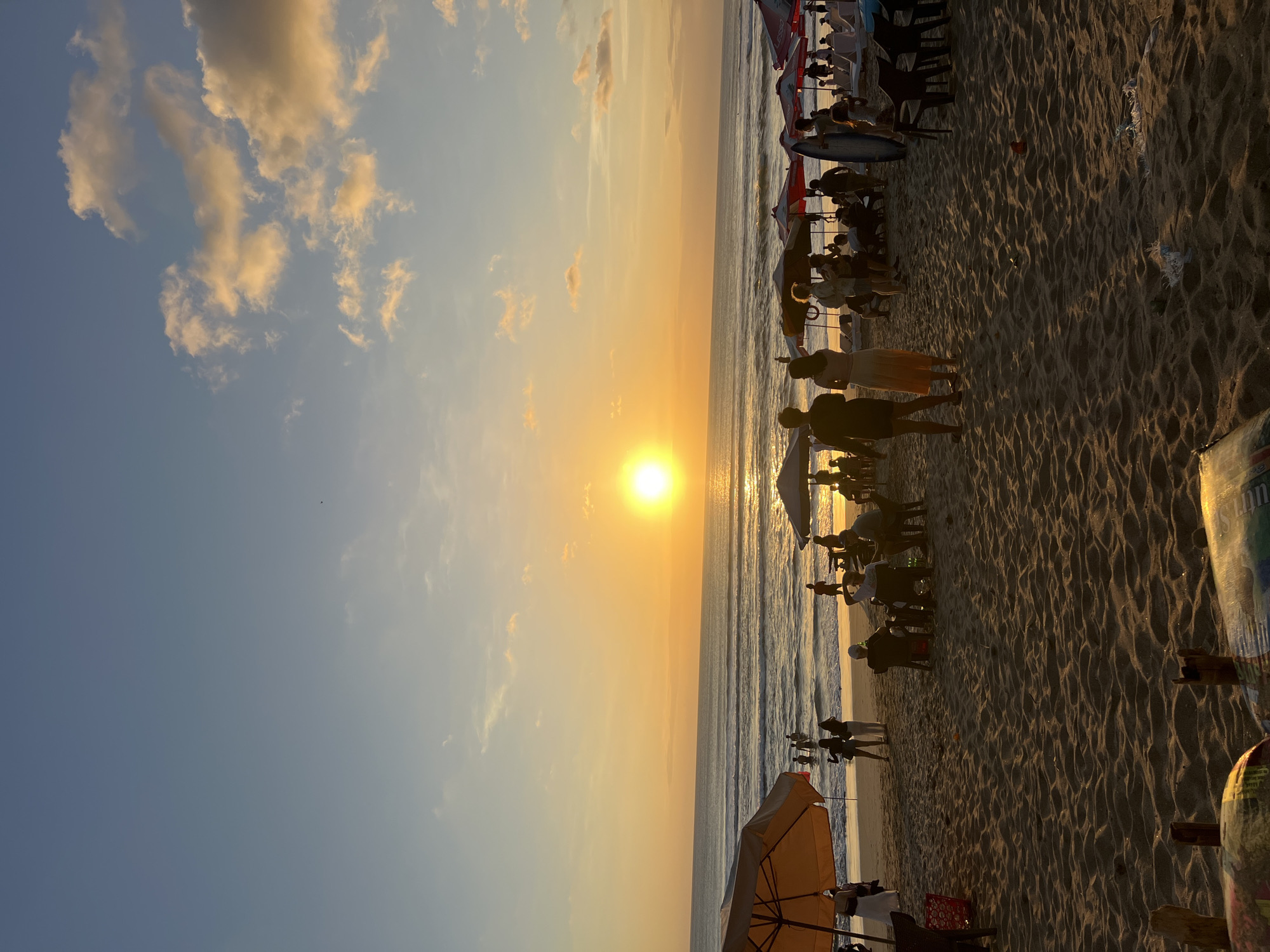 This is a low-angle, full-shot, vertical, outdoor, daytime photograph of Kuta Beach in Bali, Indonesia, during sunset. The sun, a bright, glowing orb, is low on the horizon over the ocean, casting a golden, orange hue across the sky and water. The sky is a gradient of pale blue near the top, transitioning to yellow and orange near the horizon, with scattered, wispy clouds tinged with yellow.
The ocean is calm, with gentle waves rolling onto the shore. The beach is sandy, with distinct footprints and ripples on the surface. The foreground of the image is dominated by sand, with a blurred object in the very bottom left corner.
In the middle ground, there are numerous beach umbrellas, some orange and white, others red and white, providing shade. Several people are visible walking on the beach, standing, sitting, and wading in the shallow water. A few individuals are silhouetted against the setting sun. There are beach chairs set up in some areas.
Further back, along the water's edge, more people are wading and standing. The scene depicts a typical sunset on a popular beach, with a relaxed and leisurely atmosphere.