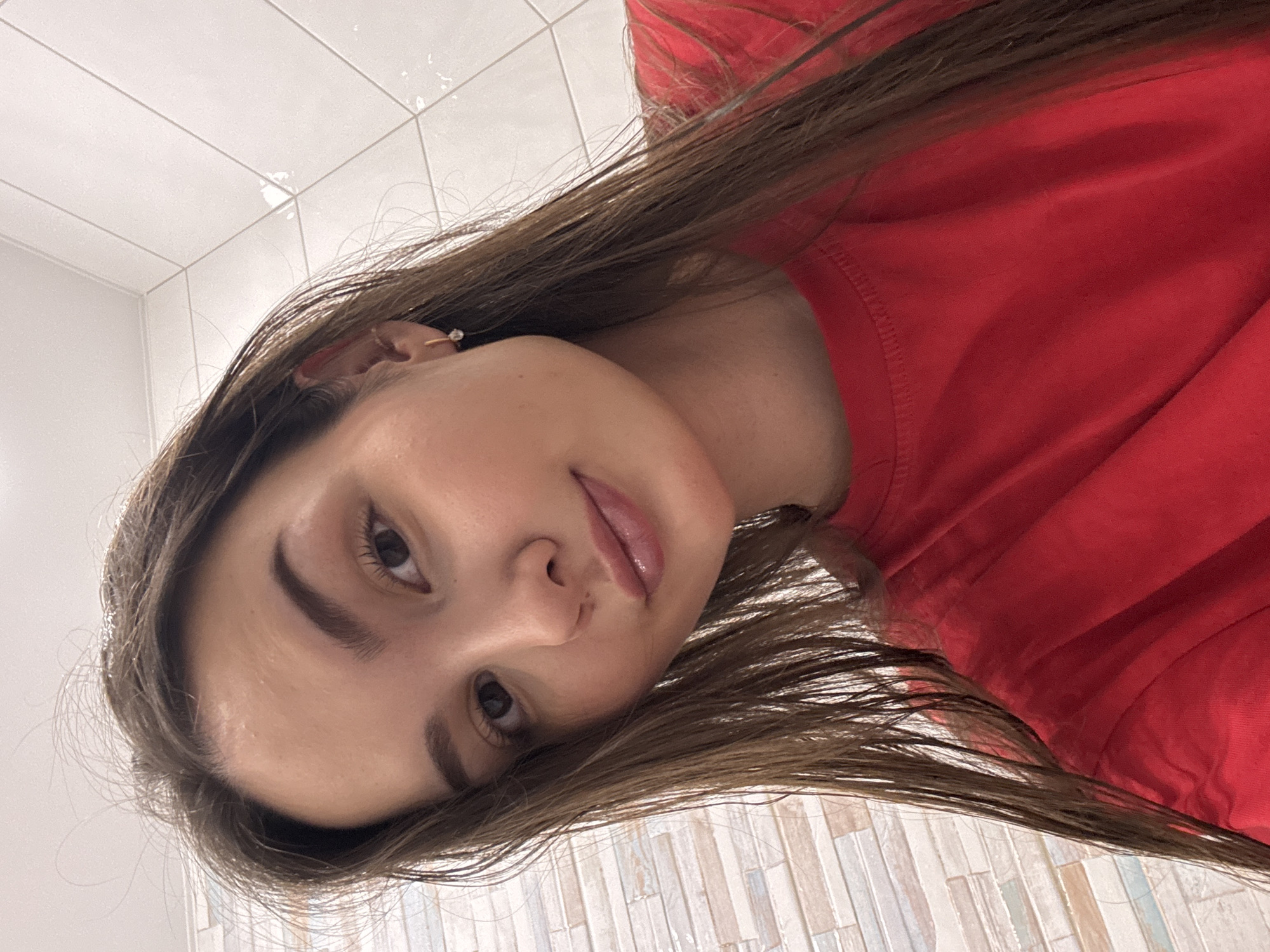 A selfie of a fair-skinned person with long brown hair, wearing a red t-shirt, indoors. The background includes white and beige tile walls. There's no indication of Chaikovsky, Russia.