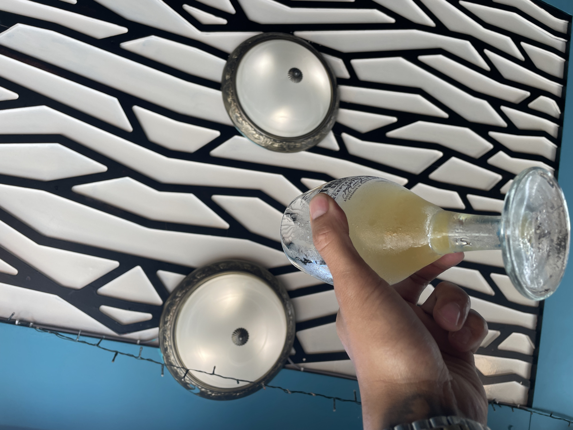 A low-angle indoor shot shows a glass containing a yellowish liquid, seemingly held by a hand and positioned against a backdrop of an elaborate ceiling design. The ceiling is characterized by a geometric pattern of black and white panels, possibly tiles, arranged in a wave-like formation. Two circular light fixtures with ornate, antique-style frames are visible, embedded within the ceiling. The foreground is dominated by the partially visible arm and wrist of the individual holding the glass, adorned with a wristwatch. A string of delicate lights is also hanging. The lower part of the frame transitions to a turquoise-blue wall. The photograph lacks identifiable landmarks to determine its location in Barcelona, Spain.