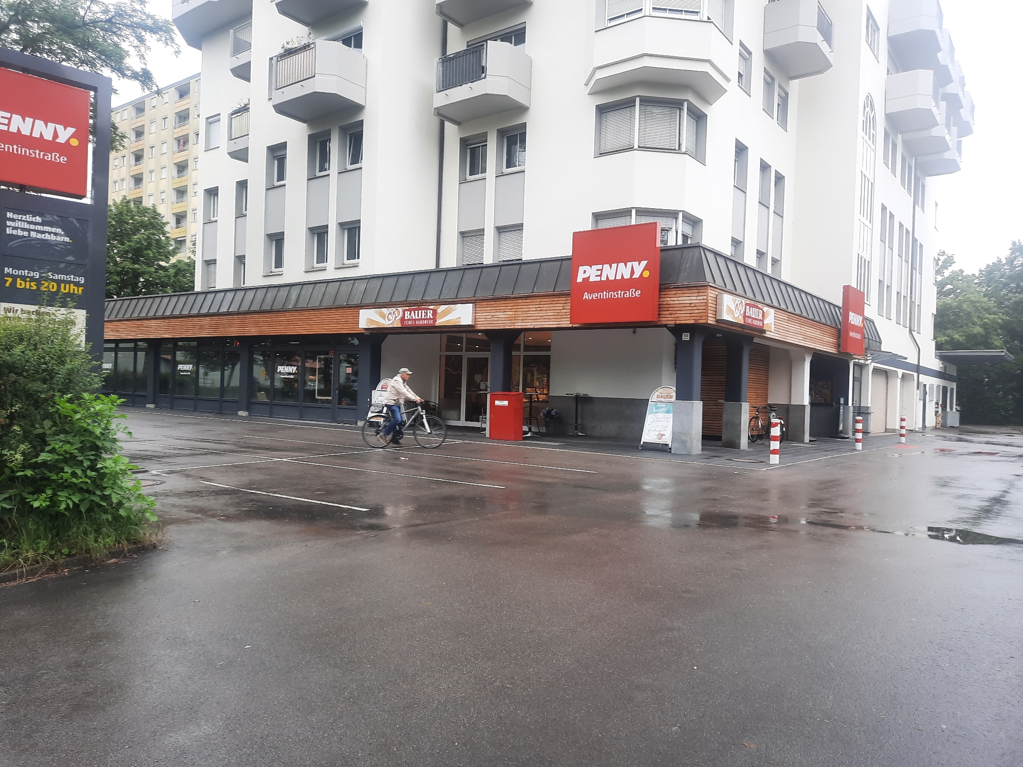 A horizontal, medium shot captures a Penny supermarket with a "Bauer Feines Handwerk" (Bauer Fine Craftsmanship) bakery section in the foreground, situated in front of a multi-story building. The scene is likely in a city, given the urban architecture. The parking lot in front of the supermarket is wet, suggesting recent rain, with visible white lane markings.
On the left side of the image, a tall, dark sign displays "PENNY." in red text, along with "Herzlich willkommen, liebe Nachbarn." (Welcome, dear neighbors.) and the opening hours "Montag - Samstag 7 bis 20 Uhr" (Monday - Saturday 7 AM to 8 PM). Further down, it reads "wir backen..." (we bake...).
A person on a bicycle is moving from left to right across the frame in the middle ground, in front of the supermarket entrance. They are wearing a light-colored jacket and jeans, and their bicycle appears to be a standard model. To the right of the bicycle, a bright red bin is positioned.
The supermarket itself has a wooden facade above its glass storefronts. The Penny logo is visible again on a red sign mounted above the awning, displaying "PENNY. Aventinstraße". A "Bauer" sign is also visible over the bakery section.
The background features a tall, white, modern apartment building with balconies and numerous windows. Across the street, another building with a yellowish facade is partially visible, interspersed with green trees. The sky is overcast, contributing to the subdued lighting of the scene. The overall atmosphere is that of a typical suburban or urban commercial area on a damp day.