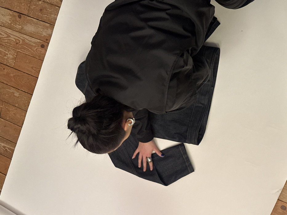 The scene depicts an interior space, identified as the city of Paris, France, configured for photography. A single individual is viewed from a high angle, leaning over a dark blue denim jacket laid flat on a white seamless backdrop. The individual's back and left profile are visible. They wear a dark, likely black, puffy jacket, and their dark hair is pulled into a bun. A silver-colored hoop earring is present on the individual's visible left ear. The left hand, adorned with blue nail polish on the thumb and index finger and a prominent silver-colored ring on the index finger, rests flat on the left sleeve of the denim jacket. The white seamless backdrop covers the majority of the visible surface, transitioning to a light brown wooden floor with visible planks along the top edge and upper left corner of the frame.