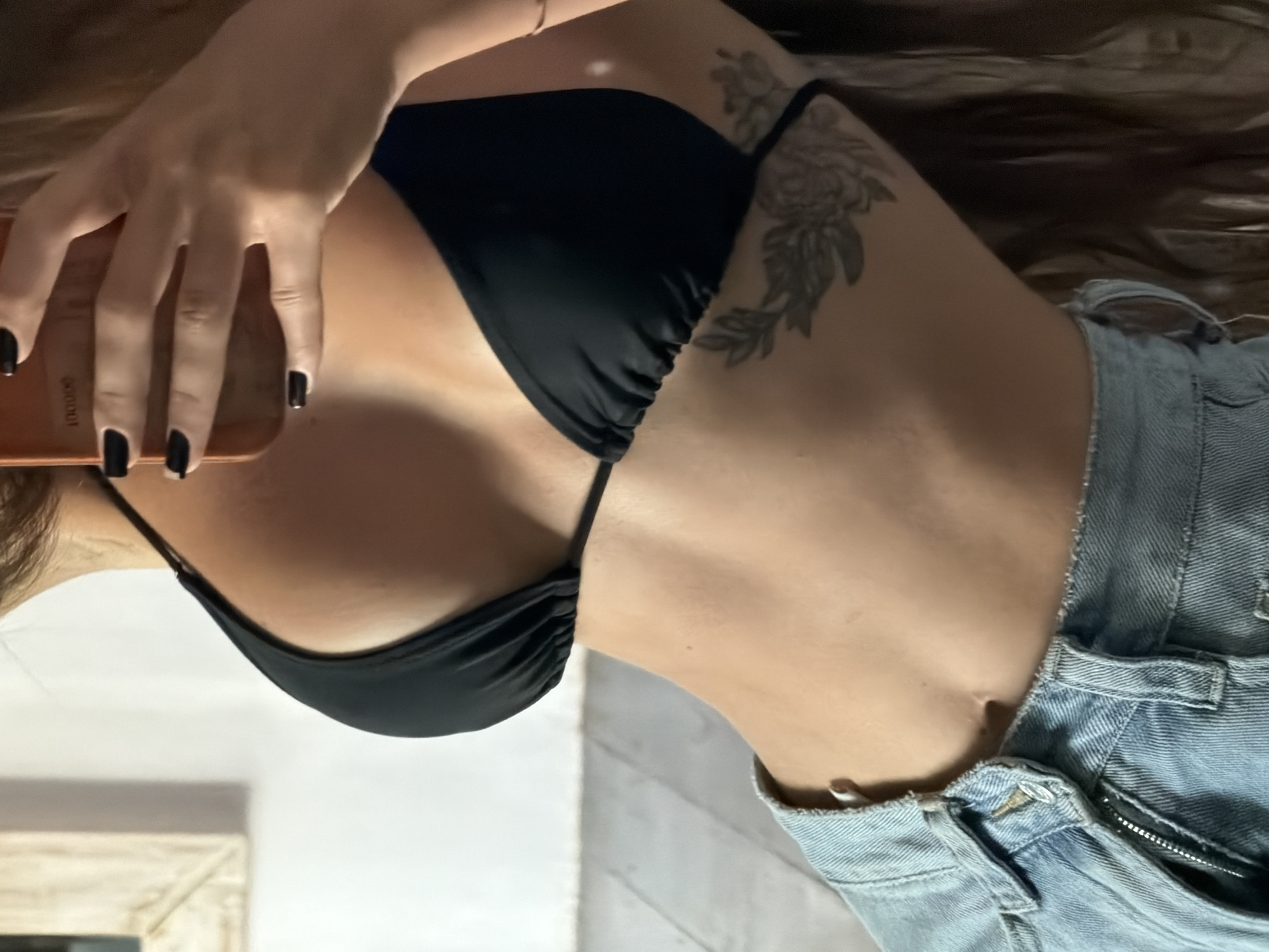 A young woman is holding a phone, taking a selfie of her upper body. She is wearing a black bikini top and denim jacket. A floral tattoo is on her left upper arm. The location is Unknown, Indonesia.