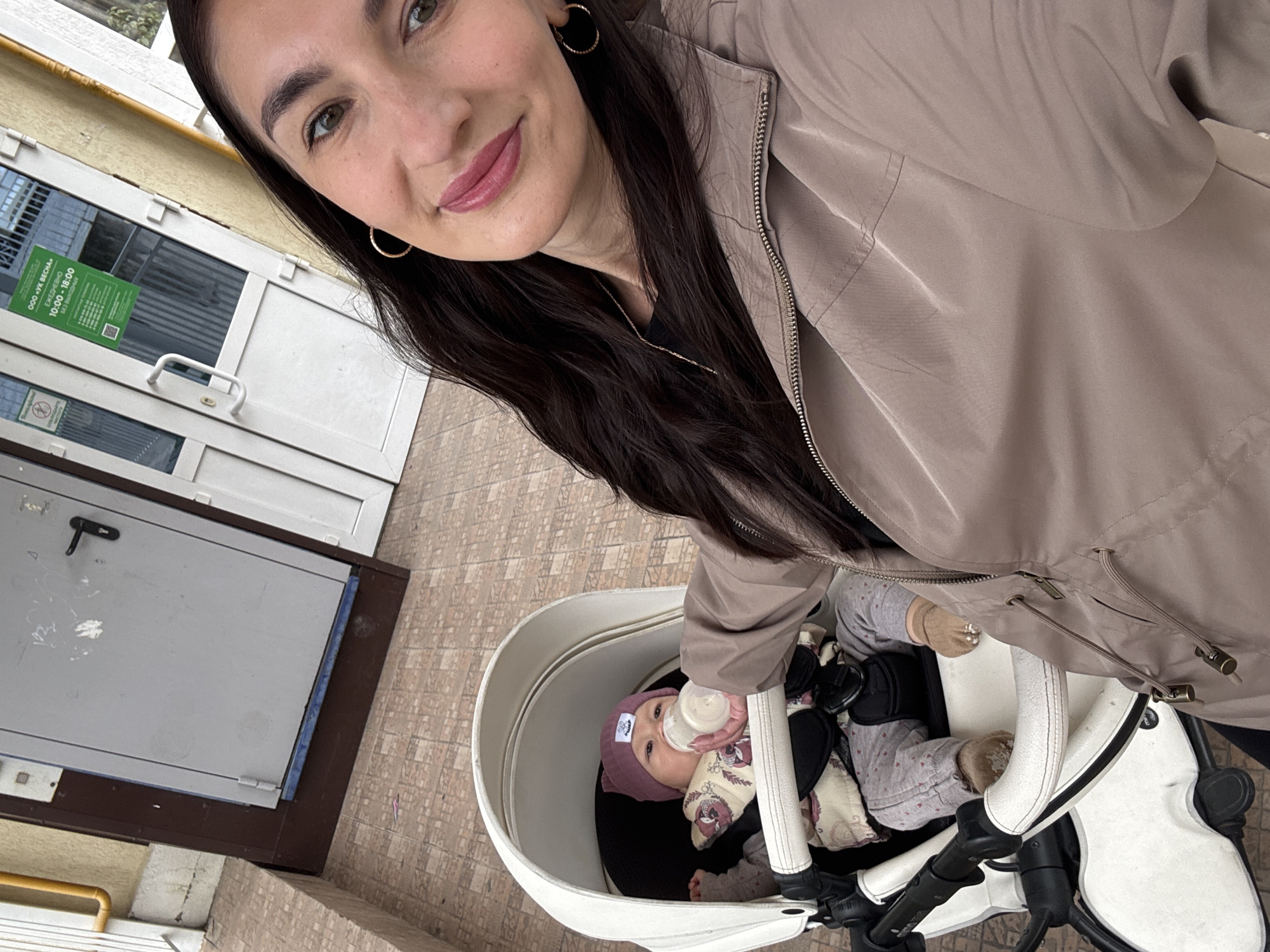 A medium-angle indoor shot depicts a woman with long, dark hair, wearing gold hoop earrings and a light brown jacket. She is holding a baby in a white stroller. The baby is wearing a purple hat and is being bottle-fed. A door with a green sign (written in Russian, but indicating the name "OOO VK Vesna" and hours of operation from 10:00 to 18:00) is visible in the background. The location is Sochi, Russia.