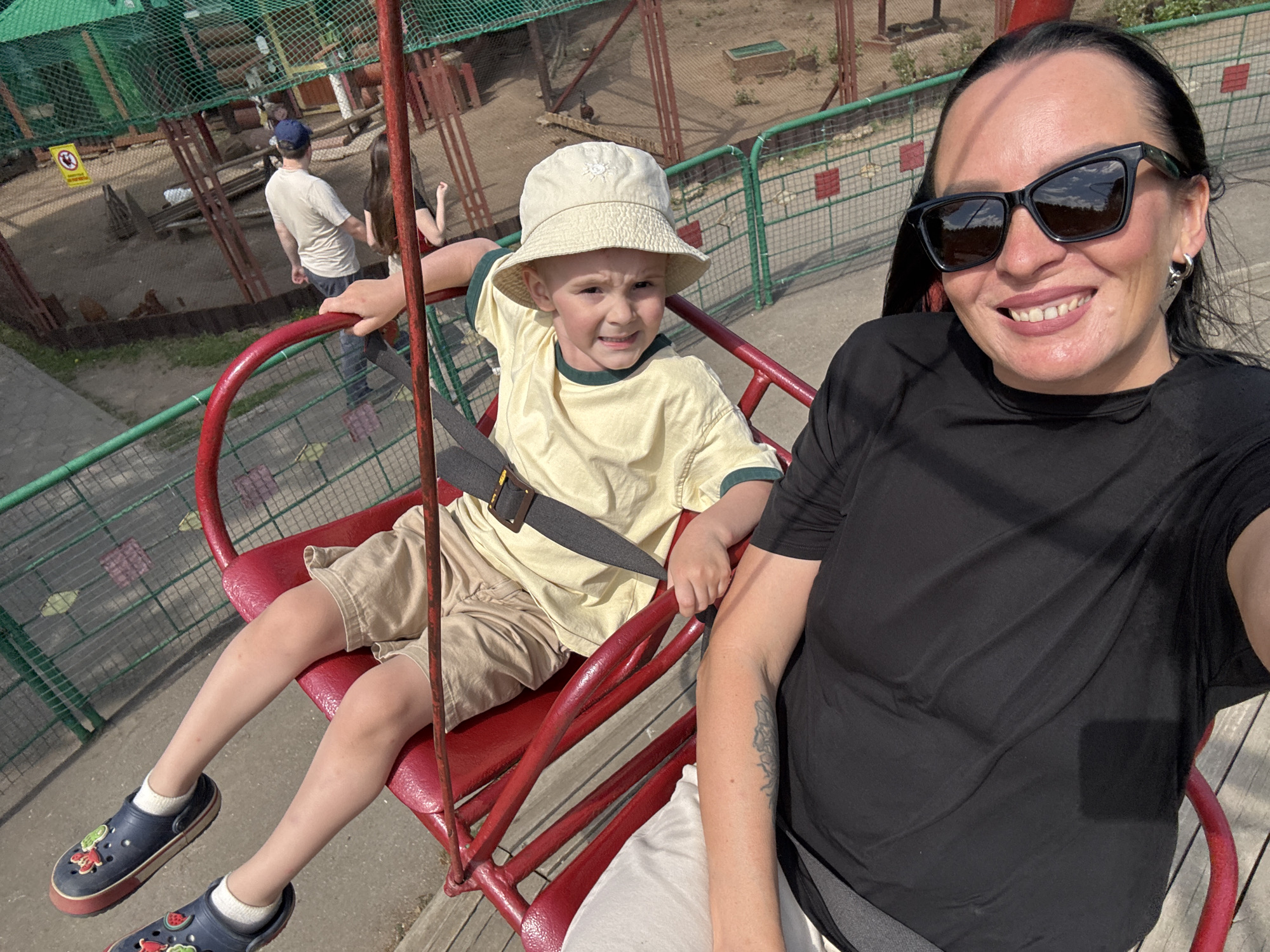 The image depicts a woman and a young boy riding a red chairlift-like ride in an outdoor setting. The boy, wearing a light yellow shirt, khaki shorts, a beige bucket hat, and blue Crocs, is securely fastened with a gray safety belt. The woman, wearing a black t-shirt and sunglasses, is smiling and taking a selfie. A man in a white shirt and a baseball cap is walking in the background, along with another person. The ride appears to be part of a zoo or park, with a green fence visible behind the riders. The location is identified as the city of Chaikovsky, Russia.