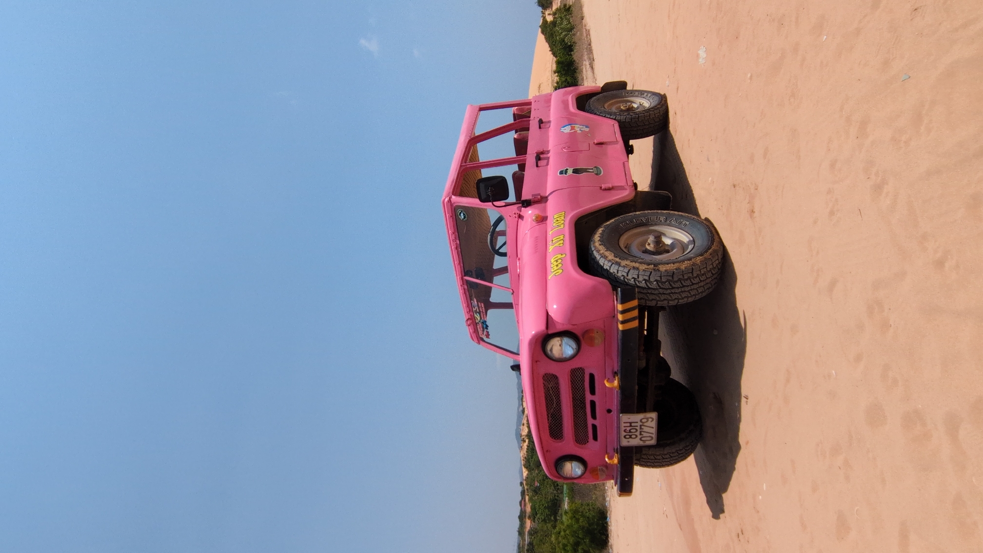 A pink, roofless off-road vehicle is parked on a sand dune in Phuoc Dinh, Vietnam. The vehicle has visible license plate "86H 0779" and writing on its side in yellow font. The location is sandy with sparse vegetation in the distance. The sky is clear and blue.