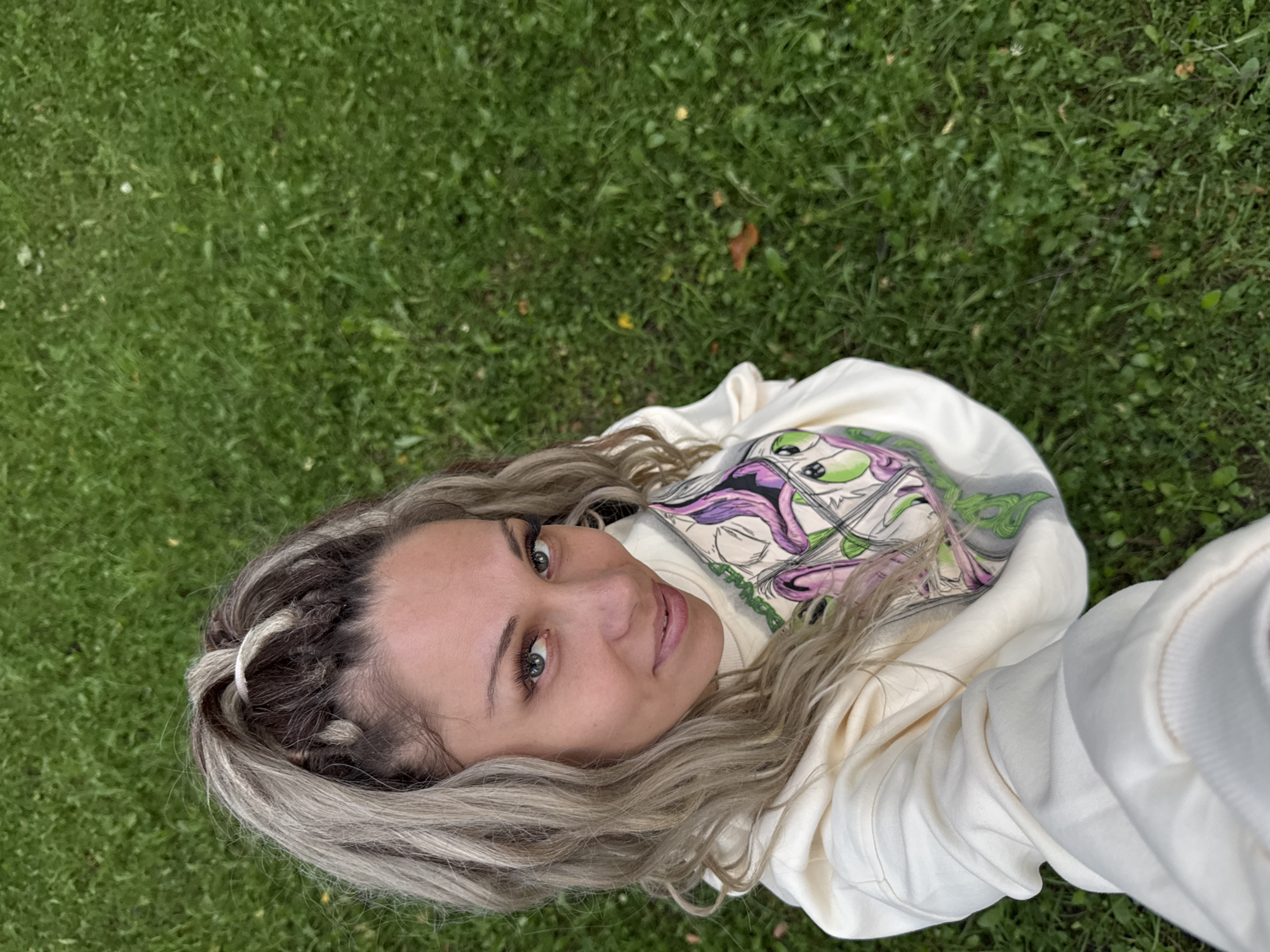 An overhead shot captures a young woman posing for a selfie on a grassy lawn in Rusanovo, Russia. She has blonde and brown braided hair pulled into a high ponytail with a white hair tie. Her eyes are green and she is smiling. She is wearing a cream-colored hoodie with a graphic print of cartoon-like characters with large eyes on the front. The background is a lush green lawn with scattered leaves and small yellow flowers.