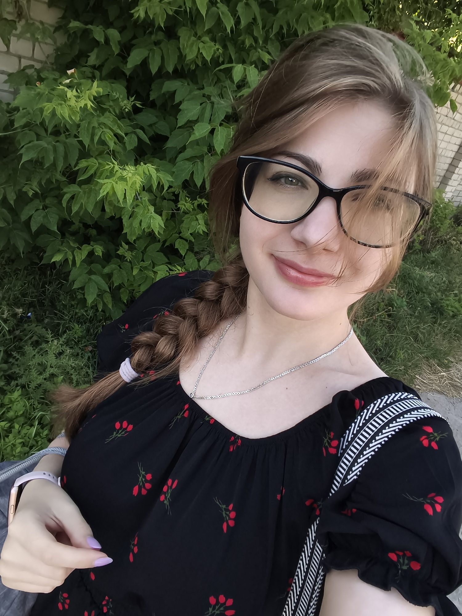 A medium shot captures a young woman with light brown hair styled in a braid, wearing glasses and a black top with a floral pattern of red berries. She is outdoors, with lush green foliage and a brick wall visible in the background. The lighting suggests it is daytime, likely sunny, as indicated by the bright highlights and shadows. The woman is looking at the camera and smiling, with a relaxed and cheerful expression. Her makeup includes subtle lip gloss and what appears to be dark eyeliner. She is wearing a silver necklace and a light-colored watch on her left wrist. The setting appears to be a natural environment, possibly a park or garden, with the focus on the woman. No text is visible in the image.