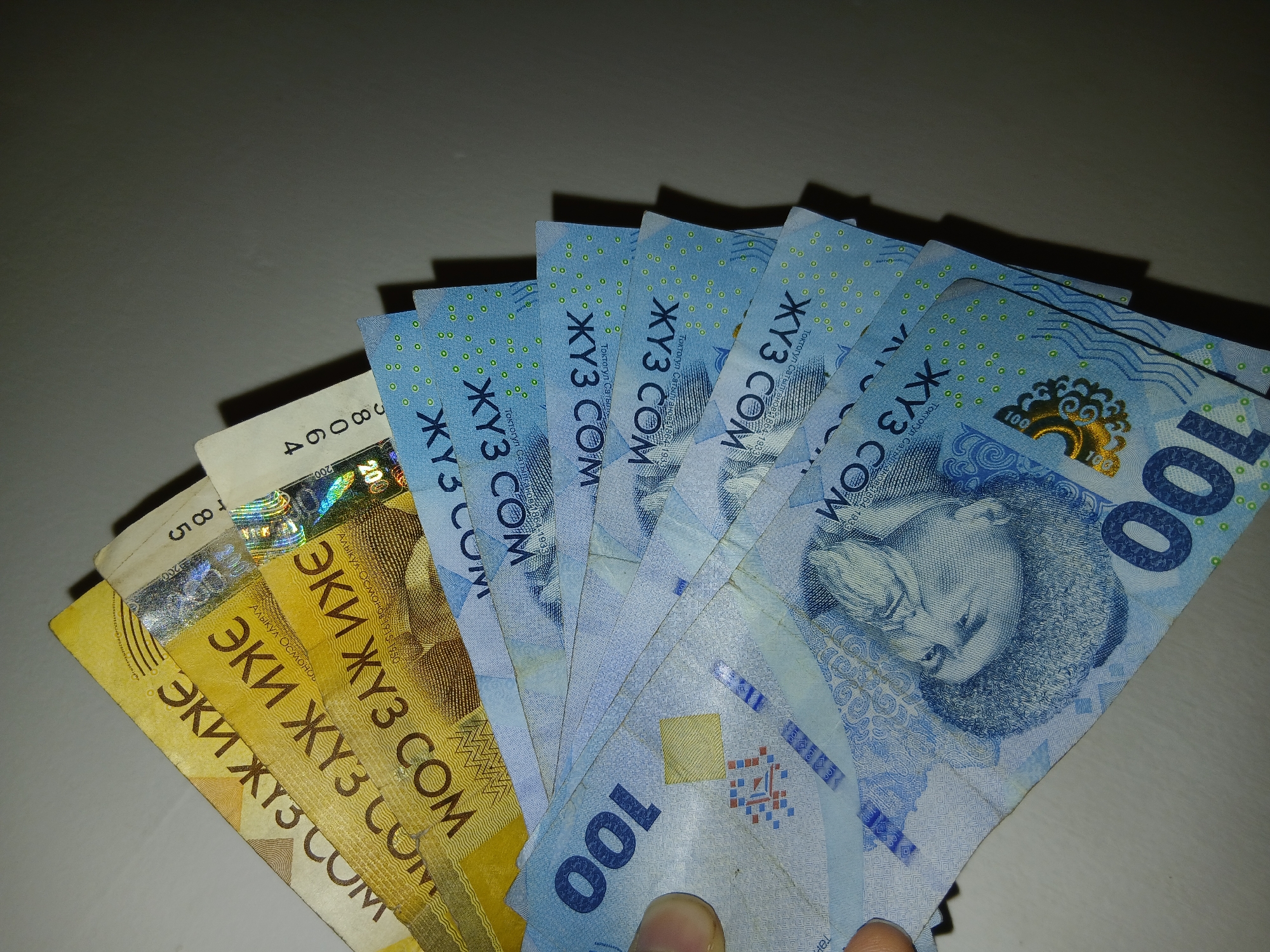 A fan of Kazakhstani Tenge banknotes is spread out on a surface. The top banknotes are yellow and white with Cyrillic text and a holographic stripe. The banknotes below are light blue and feature the number "100" in large font, a portrait of a person, and intricate patterns. The image is taken from a top-down perspective with the banknotes fanned out to the right, obscuring the majority of the background which is a plain wall.