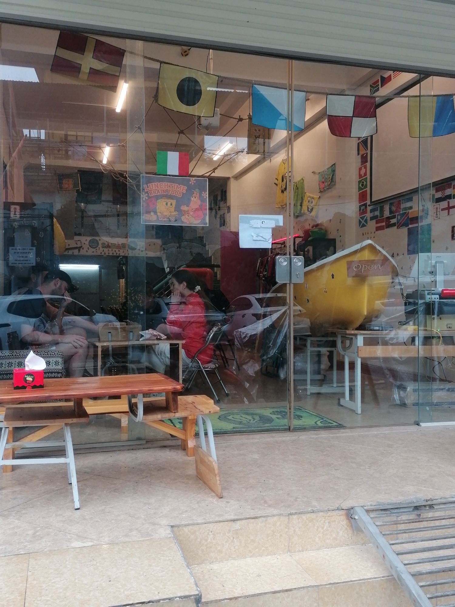 The image shows the interior of a storefront in Sa Pa Ward, Vietnam, visible through a glass window. Inside, two people sit at a small table, seemingly engaged in conversation. The man on the left wears a dark shirt and cap, while the woman on the right is dressed in a red shirt. On the table in front of them is a tissue box. In the background, behind the people, is a yellow bathtub that has the word "Open" written on it. The walls of the store are decorated with international flags, nautical pennants, and other decorative items, including a "Lunchtime Brainfood" SpongeBob SquarePants poster. Outside the window, there is a wooden table with two benches and an aluminum ramp. The ground is tiled.