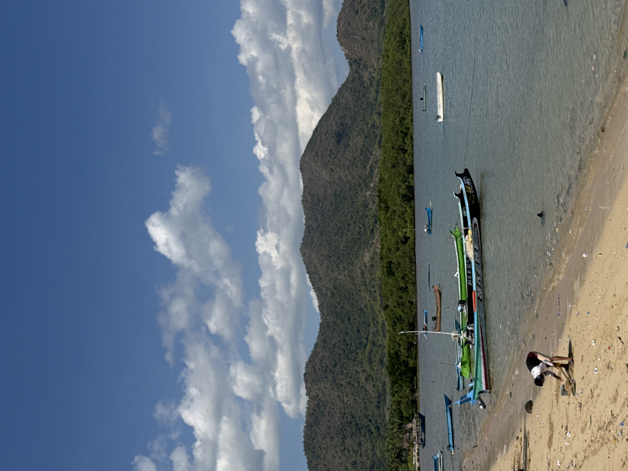 In the city of Kertasari, Indonesia, a person is seen raking a sandy beach. The beach is littered with various types of debris, including plastic bottles and other refuse. Several boats are moored in the calm, blue water. In the background, lush green trees cover a chain of mountains under a clear blue sky dotted with white clouds. The scene depicts a serene coastal environment marred by pollution.