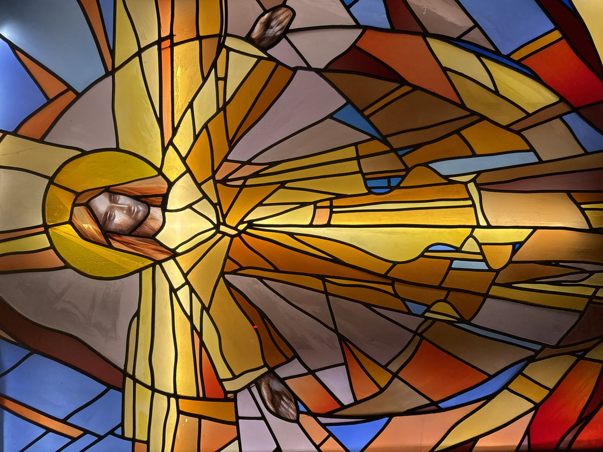 The image shows a stained-glass window, potentially depicting a religious figure, possibly Jesus. The face is centered and surrounded by a golden halo. Abstract shapes and colors, including blues, yellows, oranges, browns, reds, and grays, are arranged in a mosaic pattern that gives the impression of light emanating from the central figure. Two hands are visible on either side of the figure. The geometric shapes are outlined in black. The city and location within it are unknown.