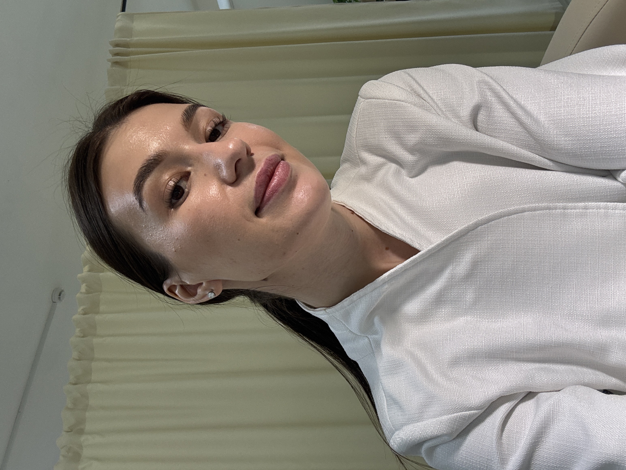 A close-up portrait shows a young woman with dark hair, dark eyes, and fair skin, smiling gently at the camera. Her head is tilted, giving the impression she is looking up towards the viewer. Her dark hair is pulled back, revealing a small silver stud earring in her visible ear. She wears a white, textured blazer or jacket, which drapes across the lower and right side of the frame. Her skin has a luminous, dewy appearance, and her lips are painted with a soft pink color.
The background is simple, featuring light-colored, pleated blinds or curtains that run horizontally behind her, suggesting an indoor setting. To the far left, a plain, light-toned wall is visible. The lighting is soft and even, highlighting her facial features without harsh shadows, implying natural light. The overall mood of the image is calm and pleasant. There is no visible text. This indoor scene is located in Chaikovsky, Russia.