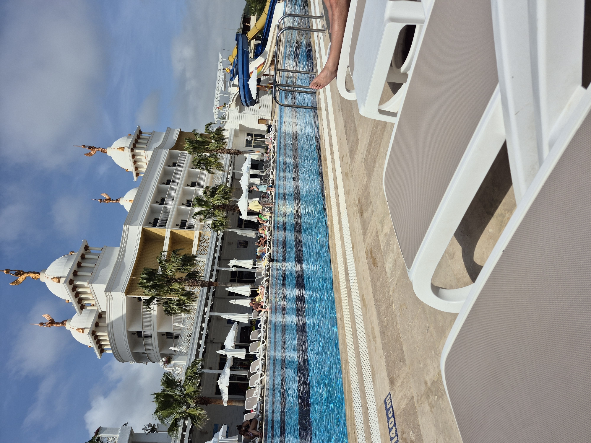 The image shows a pool area at a resort in Alanya, Turkey. A white building with domes and statues is on the left. Palm trees and white umbrellas surround the pool. Lounge chairs line the side of the pool. People are swimming and lounging around the pool. A blue water slide is partially visible in the background. A person's foot is visible near the pool's edge. The pool's depth is marked as "140cm."