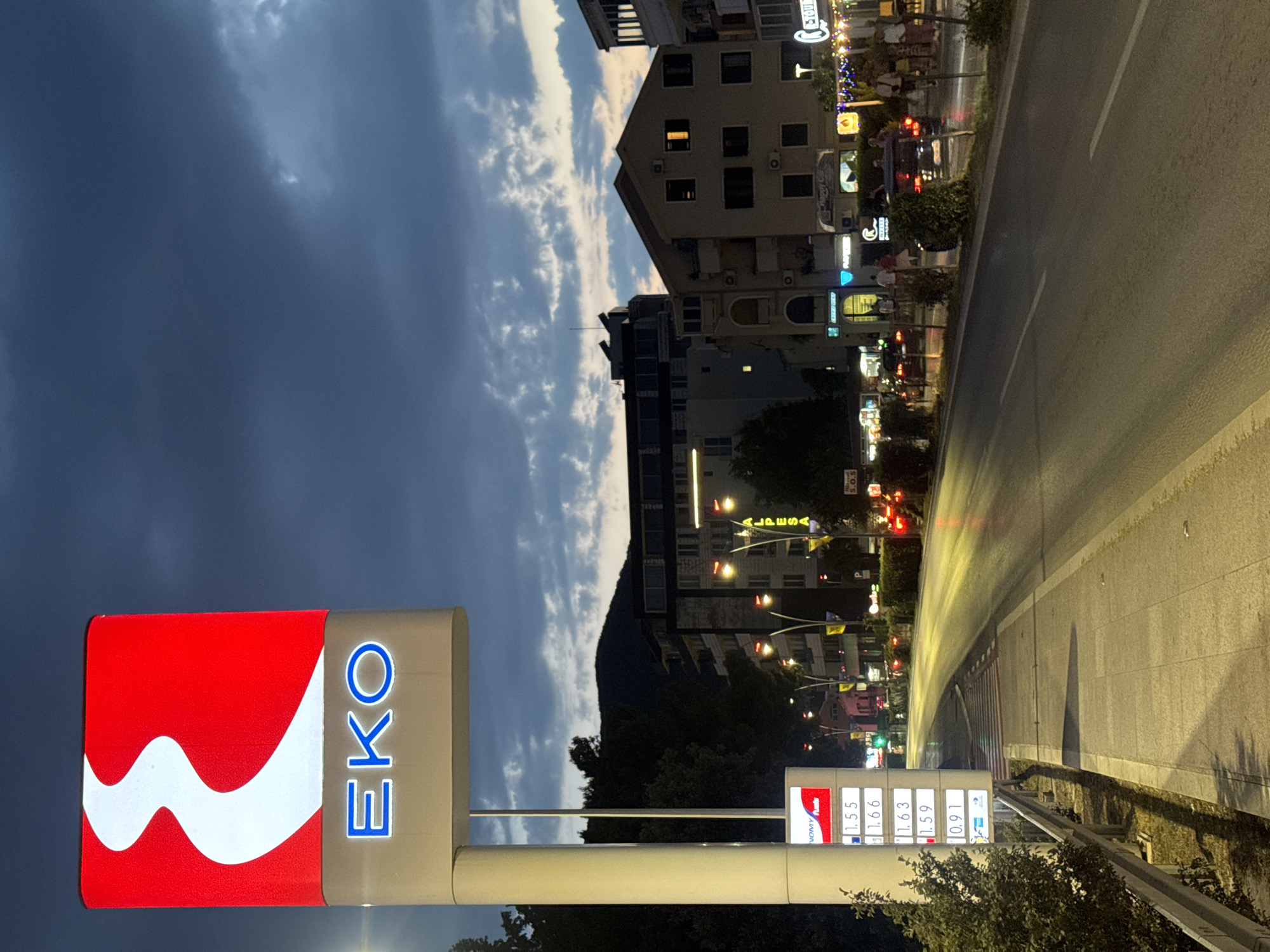 This is a vertically oriented photograph taken at dusk in Budva, Montenegro. The scene depicts a street lined with buildings, a road with a moving vehicle creating light streaks, and a gas station sign.
The primary subjects are the buildings, the gas station sign, and the road. The buildings appear to be commercial establishments or residences, with several floors and illuminated windows. The gas station sign, prominently displayed on the left, features the "EKO" logo in blue letters against a brown background, with a large red and white graphic. Below the main sign, a price board displays fuel prices: 1.55, 1.66, 1.63, 1.59, and 0.91, with an "OMV MaxxMotion" logo.
The activity taking place is a street scene at the end of the day. The moving vehicle on the road suggests traffic, and the illuminated buildings and streetlights indicate that it is nighttime or late evening. The sky is dark blue with some lighter clouds, characteristic of dusk.
Notable details include the lighting, which creates a vibrant atmosphere with streetlights and building illumination. The light streaks from the moving vehicle add a dynamic element to the image. The presence of the "EKO" and "OMV MaxxMotion" logos clearly identify the location as a gas station. The mountain in the background, partially obscured by trees, provides a sense of the natural landscape surrounding the urban area. The overall mood is one of evening activity in a town or city.