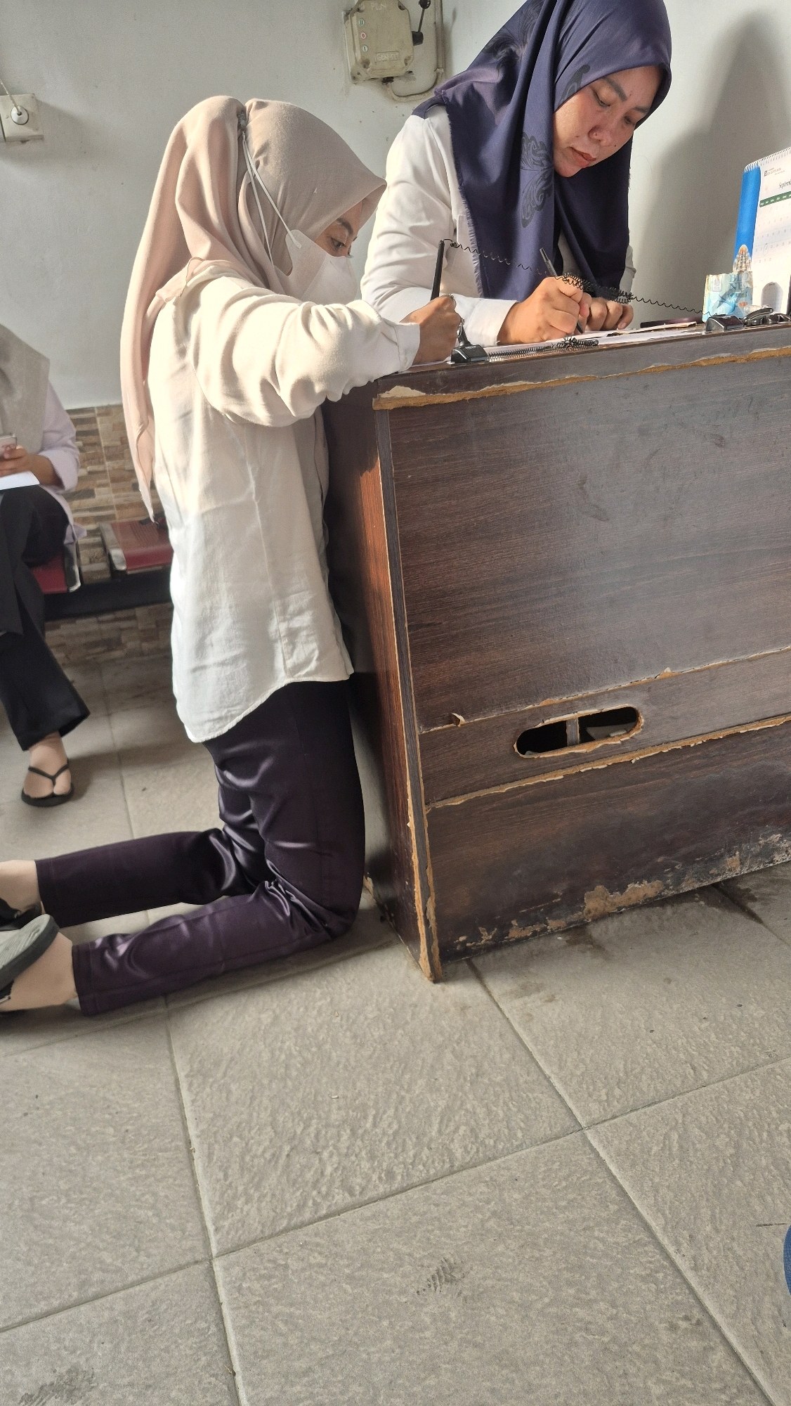 Two women are kneeling at a wooden counter in what appears to be an office or administrative setting. The woman on the left, wearing a light-colored hijab and white face mask, is writing on a document with a pen. The woman on the right, wearing a dark blue hijab and white shirt, is also writing on a document. They are both facing a desk with papers and a calendar on it. The floor is tiled. The scene suggests an indoor administrative process in Jambi, Indonesia.