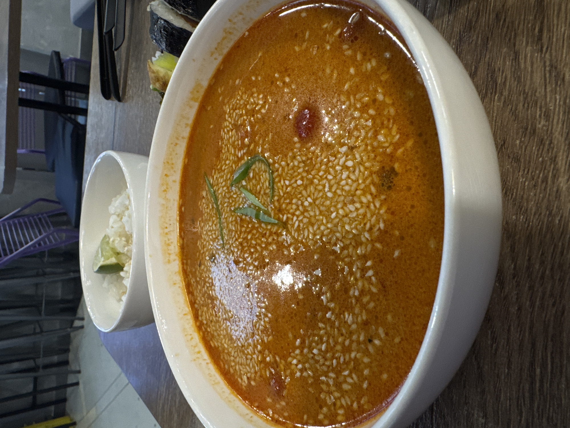 A close-up shot of a bowl of soup, possibly Tom Yum soup, is displayed next to a smaller bowl of rice on a wooden surface. The soup is a rich orange color, garnished with green herbs and sprinkled with sesame seeds. The smaller bowl contains white rice, with a slice of lime visible. In the background, cutlery, sushi rolls, and a partially visible chair with a purple frame are present. The location is potentially Mozhvai, Russia.