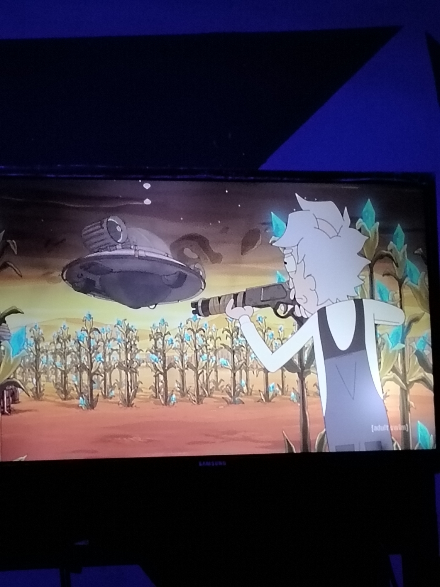A television screen displays an animated scene from Rick and Morty. On the left, a silver, disc-shaped alien spacecraft hovers above a field of tall, thin plants with bright blue crystalline growths. On the right, a white-haired animated character, presumably Rick Sanchez, wearing a white shirt and grey overalls, aims a futuristic rifle. The background is a dusty, orange-brown landscape with a dark, cloudy sky. A small black bar with the word "SAMSUNG" is visible at the bottom of the screen. The scene is illuminated by the television's display, casting a blueish glow on the surrounding environment. The television is positioned in a dark room, with a dark blue, angled surface visible at the top. The location in Du, Nigeria, is not discernible from the image.