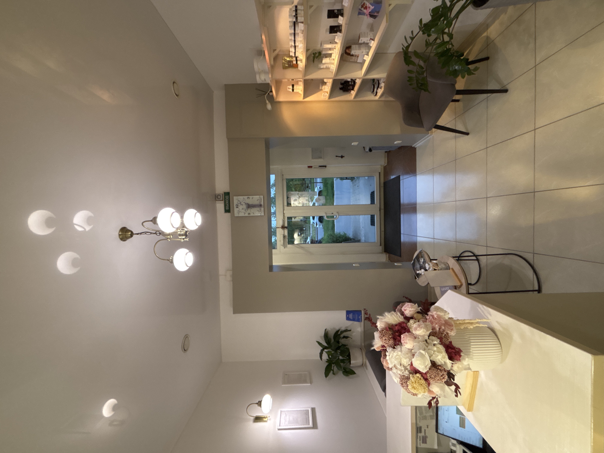 The image depicts the interior of a lobby or reception area, possibly in a spa or salon setting, located in an unspecified urban location. The space is well-lit and features a modern aesthetic.
The focus is drawn to the reception desk, which is white with a prominent display of artificial flowers in a white vase. Behind the desk, a computer screen is visible. To the left, decorative wall lamps are present, along with a potted plant.
Beyond the reception area, there is a doorway leading to an exterior space. Through the doorway, one can discern outdoor scenery.
On the right side of the image, a shelf unit displays various products. Adjacent to the shelves, a chair is positioned, accompanied by another potted plant.
In the upper portion of the image, a chandelier-style light fixture is affixed to the ceiling, providing illumination to the area.