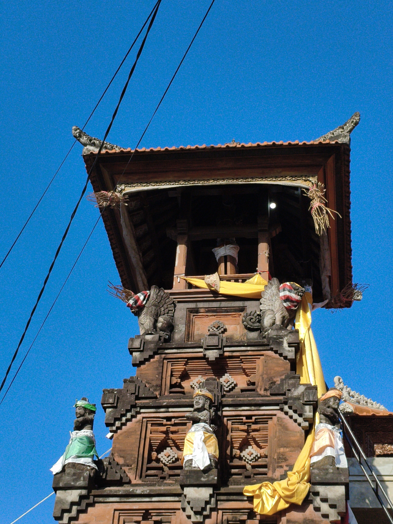 This is a low-angle, medium shot of the Bale Gajah temple in Denpasar, Indonesia, taken against a clear blue sky. The temple is constructed of red brick and features ornate carvings and decorative elements. At the top is a tiered roof supported by wooden beams, with a yellow cloth draped over the side. Several statues adorn the temple's structure, some wearing traditional Balinese clothing. Two winged mythical creatures are carved on either side of the upper section, and a third carved figure is visible on the left side. The temple is partially obscured by black electrical wires running diagonally across the frame. The overall scene depicts a religious structure with intricate architectural details.