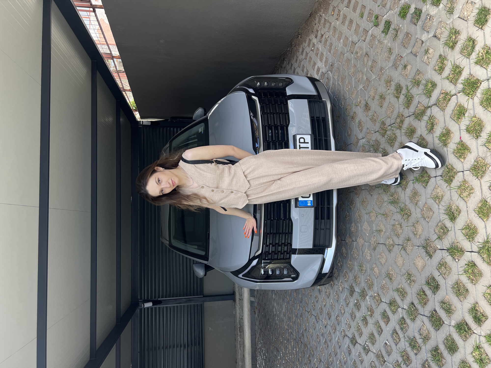A woman with long brown hair is lying on the hood of a silver car. She is wearing a light brown jumpsuit and white sneakers with black accents. The car is parked in what appears to be a driveway or garage with a concrete block pattern floor and a metal structure overhead. A license plate is visible on the front of the car. The location is Unknown, Unknown.