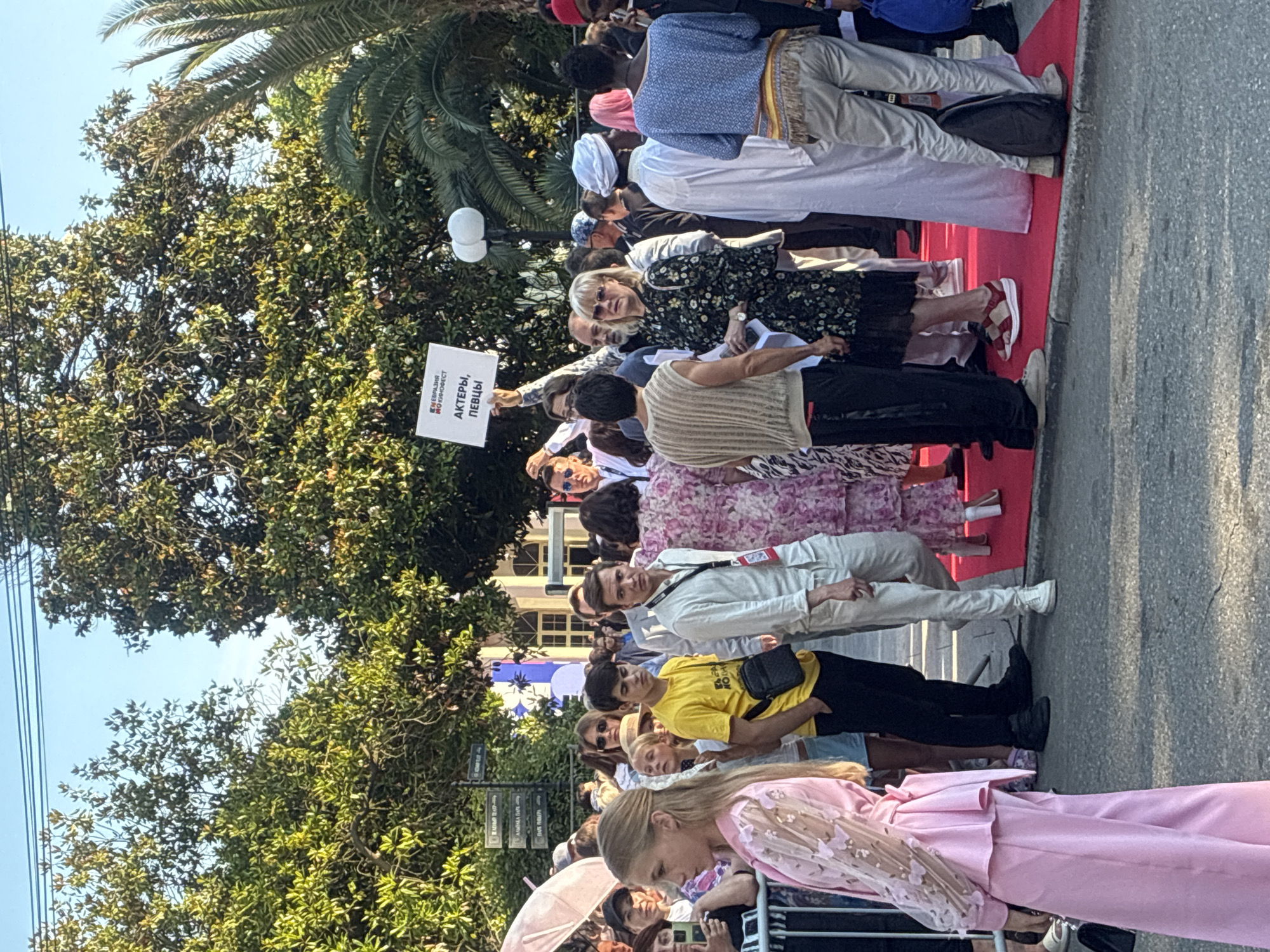 This is a vertical, eye-level, medium shot of a crowd of people on a red carpet on a sunny day, likely in Sochi, Russia, given the foliage and palm trees. The people are dressed in a variety of casual and formal attire, including a woman in a floral dress, a man in a white suit, and a woman in a pink outfit. Several individuals are holding signs, one of which is clearly visible and reads "АКТЕРЫ, ПЕВЦЫ" (Actors, Singers) in Russian. The presence of a red carpet and the signs suggest an event or premiere. The atmosphere appears festive and crowded, with people looking towards the camera or conversing amongst themselves. The time of day is likely afternoon, indicated by the bright sunlight and shadows.