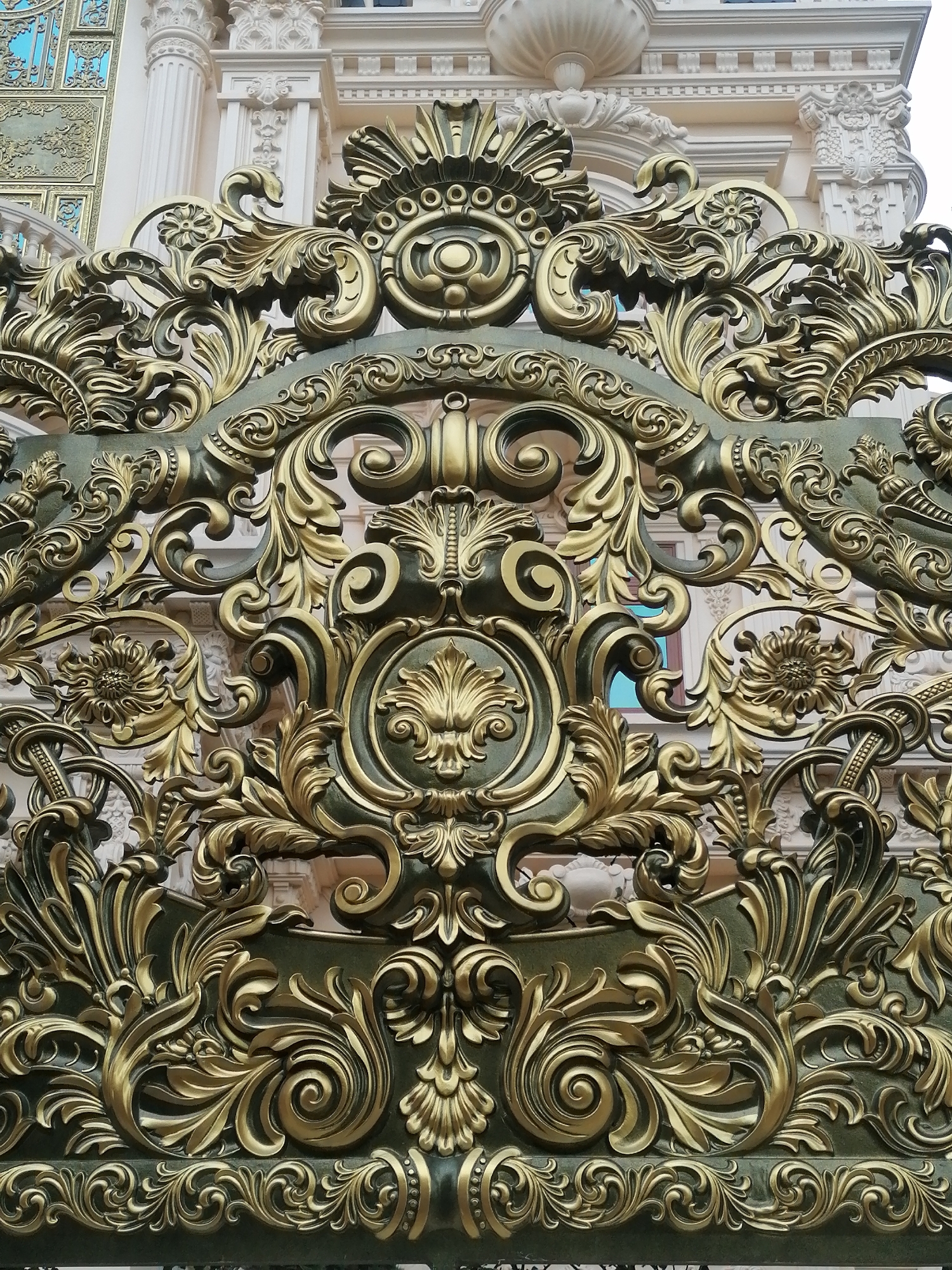 This close-up image features an ornate, gilded metal gate or railing, rendered in a dark bronze or deep green base color with intricate details highlighted in a bright gold finish. The gate is elaborately decorated with swirling scrollwork, volutes, and highly detailed floral motifs, including acanthus leaves and stylized flowers, arranged in symmetrical patterns.
In the background, parts of a grand, light-colored building with classical architectural elements are visible. These include fluted columns, ornate capitals, detailed cornices, and decorative carvings. Some sections of the building also show light blue details, possibly windowpanes or decorative inserts, contrasting with the building's cream or white facade.
The scene is static, focusing entirely on the architectural and decorative craftsmanship. The lighting suggests it is daytime. The overall impression is one of opulence and intricate artistry, indicative of grand European-inspired architecture often found in locations like Da Lat, Vietnam. There is no visible text in the image.