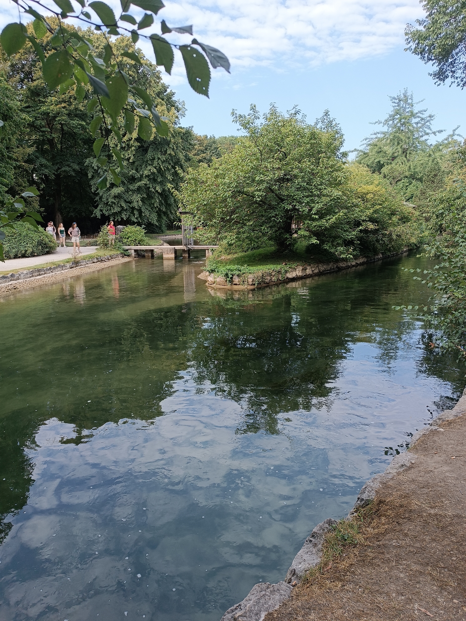 The image depicts a serene scene along a canal in Munich, Germany, likely within the Englischer Garten. On the left bank, a paved pathway is visible, bordered by lush greenery and stone walls. Several individuals are walking along this path, some appearing to be tourists. The canal itself is a prominent feature, its dark, green water reflecting the surrounding trees and the sky. A small island with dense foliage sits in the middle of the canal, and a footbridge spans the water, connecting to structures on the far bank. The environment is characterized by abundant trees, a clear blue sky with scattered clouds, and the calm flow of water. The right bank features a rougher, unpaved edge with grass and stones.