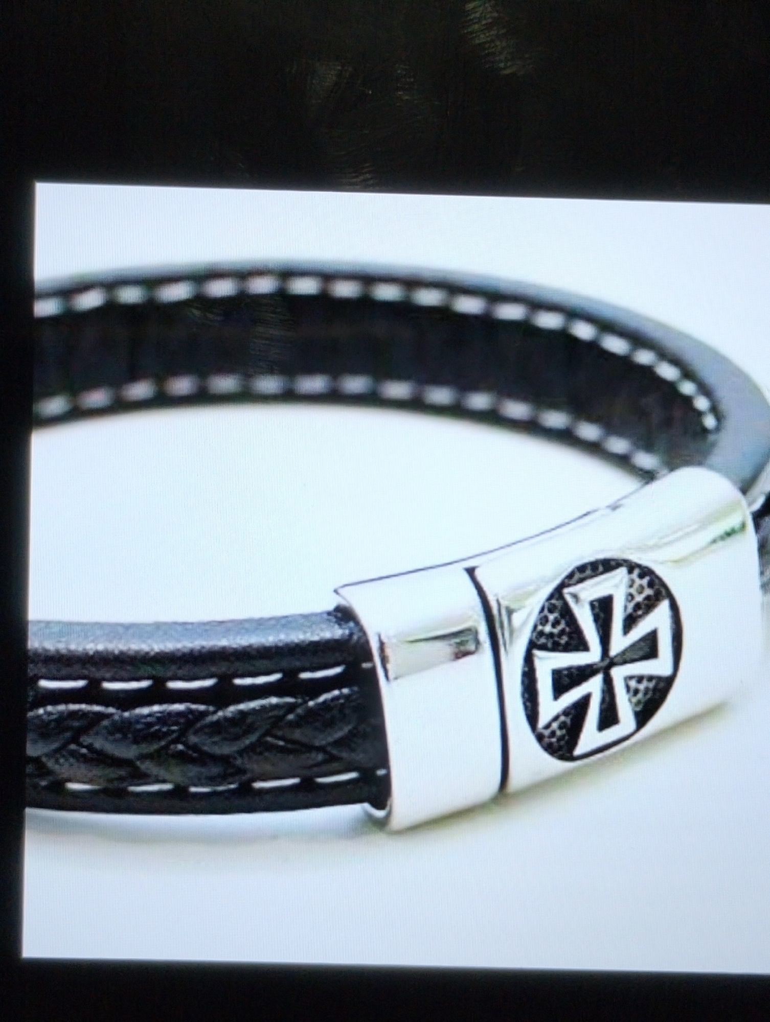 The image shows a close-up of a black leather braided bracelet with white stitching. The bracelet has a magnetic clasp that is silver in color. The clasp is adorned with a black symbol that appears to be a cross with a textured inner circle. The background is a plain white surface. The image is taken from a slightly elevated angle, focusing on the clasp and a portion of the bracelet.