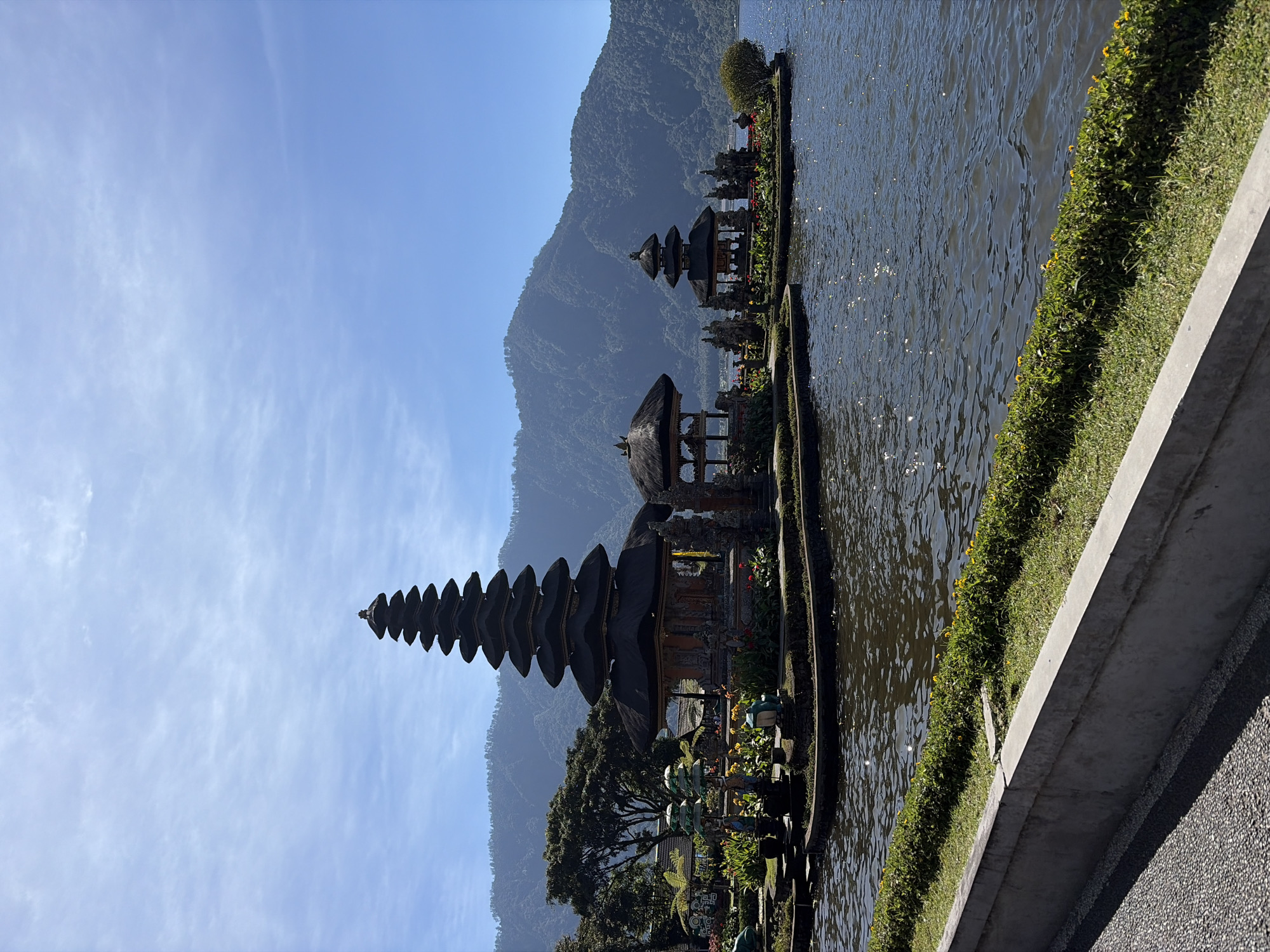 In the city of Baturiti, Indonesia, a picturesque scene unfolds at Pura Ulun Danu Bratan, a water temple situated on the shores of Lake Bratan. The image captures the main temple complex, characterized by its distinctive multi-tiered meru towers, a testament to Balinese Hindu architecture. The largest meru, a towering structure with eleven tiers, stands prominently on a small island within the lake. To its right, a smaller meru with three tiers is visible, also on its own island.
The temple structures are adorned with traditional carvings and intricate details, reflecting the rich cultural heritage of the region. Surrounding the temple on the islands are lush gardens filled with vibrant flowers and greenery, creating a serene and beautiful ambiance. The water of Lake Bratan, with sunlight glinting off its surface, forms a shimmering expanse in the foreground, reflecting the clear blue sky and the surrounding mountainous landscape.
In the background, the majestic hills of Baturiti rise, their slopes covered in verdant foliage, providing a dramatic and breathtaking backdrop to the temple. The sky above is a brilliant blue, with a few wispy clouds adding a touch of softness to the scene. The overall impression is one of tranquility, spiritual significance, and the natural beauty of Bali. There are no people visible in the image.