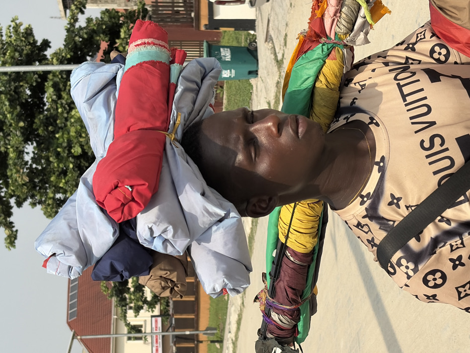 An adult male is depicted in a close-up, outdoors on a clear day in Igboefon, Nigeria. He wears a light-colored t-shirt patterned with "LOUIS VUITTON" text and monograms, and a thin gold-colored chain. A black strap crosses his right shoulder. His eyes are closed or heavily squinted, and his expression is neutral.
He carries a substantial load of merchandise: a stack of rolled fabric items, predominantly light blue/grey with a prominent red one, balanced on his head. Additionally, a long pole or bar rests on his left shoulder, from which numerous other rolled fabrics of various colors (green, yellow, maroon, brown, multi-colored) are suspended.
The background reveals a paved surface. To the left, a low orange-brown wall separates the foreground from a building with a terracotta-tiled roof, featuring visible solar panels. Green trees partially obscure the building. A white sign with red text is partially visible. To the right, a green "HAPPY FAMILY" branded refuse bin is present, alongside a partial view of a white vehicle and further vegetation. The scene is illuminated by strong sunlight, indicative of daytime.