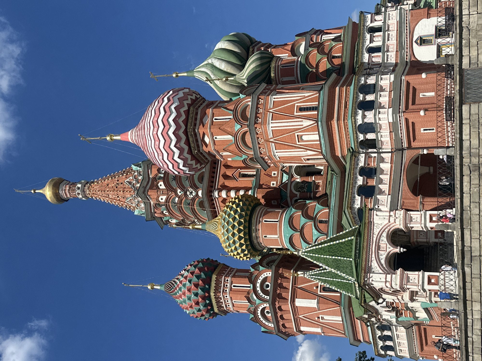 A low-angle, full shot captures the exterior of Saint Basil's Cathedral in Moscow, Russia, under a clear, bright blue sky. The iconic, colorful onion domes of the cathedral dominate the frame, with their intricate patterns and vibrant hues of red, green, gold, and pink. The cathedral's brick facade is detailed with white accents and arched windows. In the foreground, a stone wall with a black metal fence separates the viewer from the base of the cathedral. Several people are visible standing near the fence and on the cathedral's grounds, appearing to be tourists. The scene is bathed in bright sunlight, casting subtle shadows.