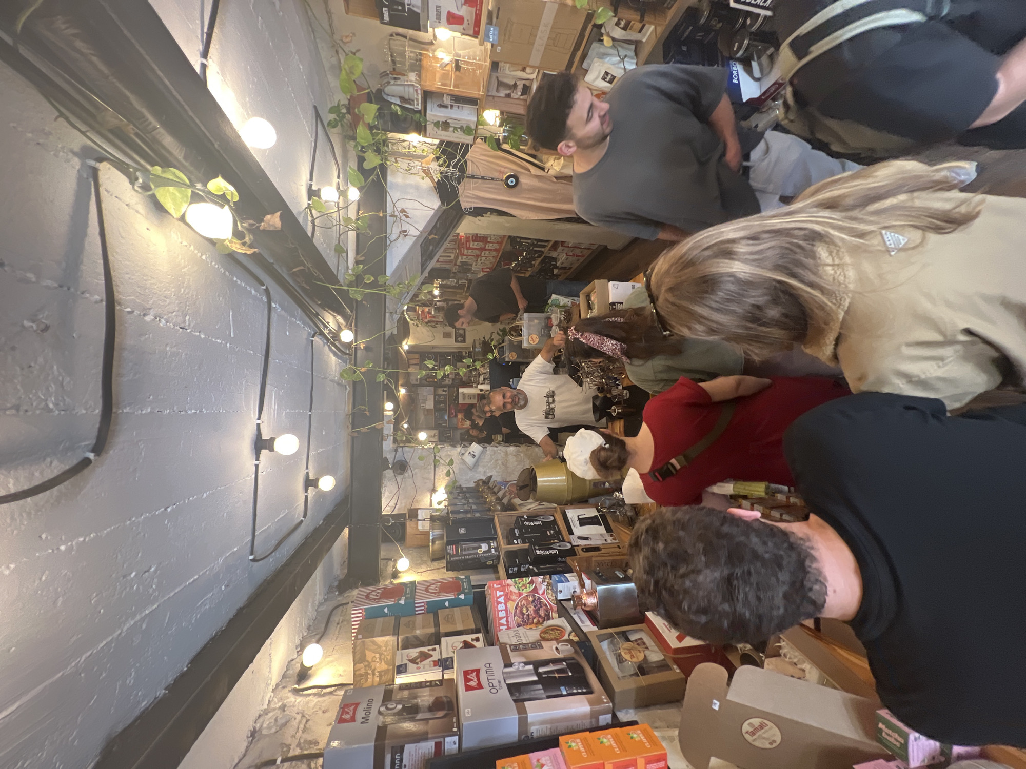 Here is a concise description of the image:
**Scene:** An interior shot of a small commercial space, possibly a retail store or cafe, seemingly located in the city of Tel-Aviv, Israel, based on the store's inventory. The focus is on the crowded interaction happening around a display counter.
**People:** A group of people is gathered around the counter. There is a man with a beard and a white shirt behind the counter, presumably a salesperson or employee. Several customers are present, including one in a red shirt, another in a black shirt, and a woman with blonde hair wearing a khaki jacket. Another person in a gray shirt stands further back.
**Objects:** The counter is covered with various boxed products, including coffee-related items like "Melitta Optima Timer," "Bialetti," and "Molino," and some other packaged goods labeled "Tamati," "Matcha," "Clipper," and "Habbat Baba."
**Actions & Interactions:** The customers are looking at and possibly interacting with the products displayed on the counter. The man behind the counter appears to be engaging with the customers, potentially offering assistance or information.
**Environment:** The store interior is decorated with string lights and greenery along the walls. The walls are painted grey. Shelves behind the customers display a variety of merchandise, creating a sense of a well-stocked, possibly small, retail space.