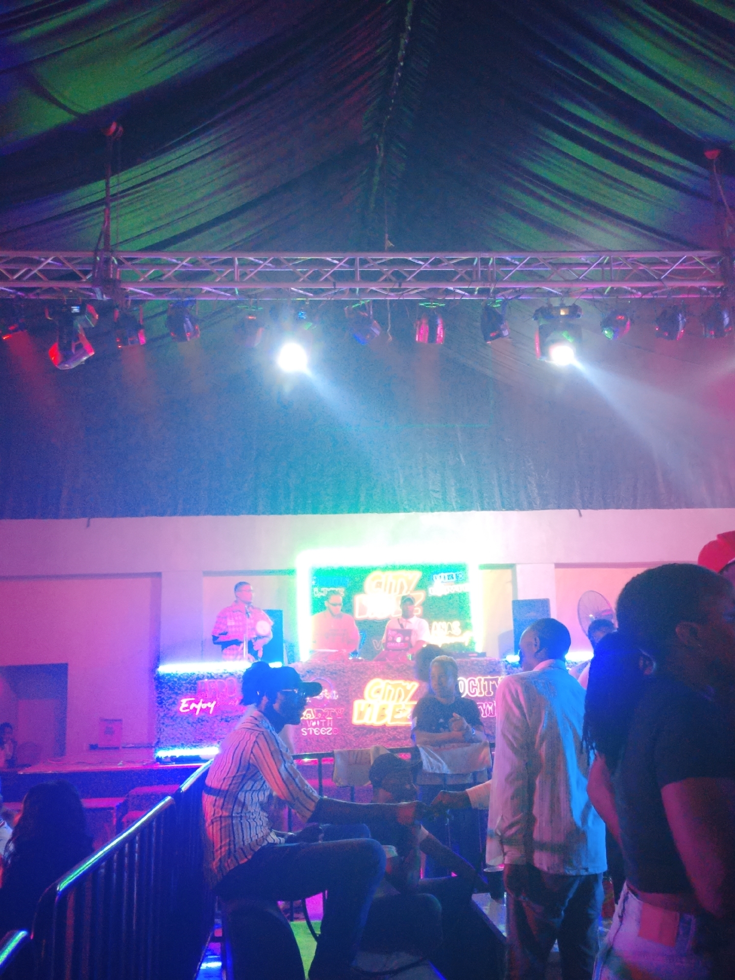 The image depicts a night scene inside what appears to be a nightclub or event space in Rido, Nigeria. The venue is illuminated with colorful stage lighting, including spotlights and neon signs. A metal truss system with attached lights is visible overhead, supporting a draped ceiling. A stage is set up with DJ equipment and neon signage reading "CITY PARTY WITH STEEZ." Several people are present, including individuals standing and seated in the foreground, and figures on stage appearing to perform or DJ. The crowd includes people of diverse dress, some in casual attire and others in more formal wear. The environment has a party atmosphere, with visual indicators like the staging and lighting suggesting a public event.